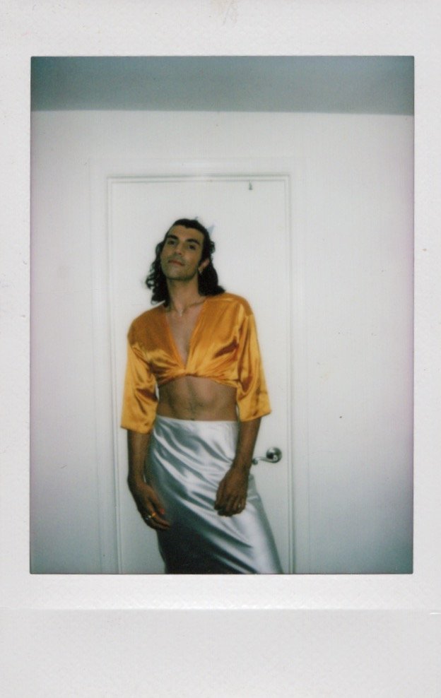 Person wearing a yellow crop top and silver skirt posing against a white wall.