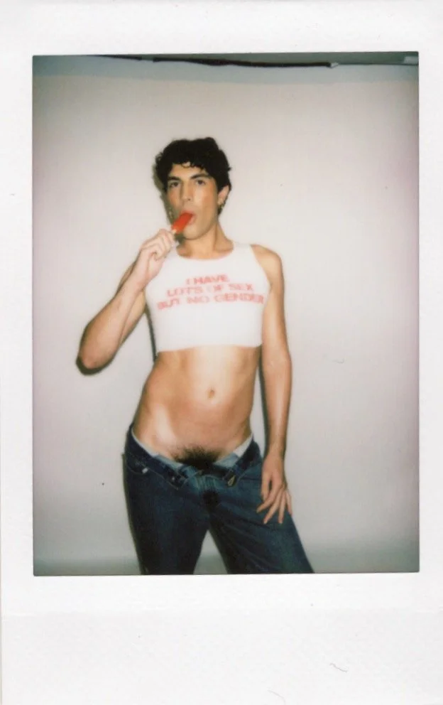 Person in a cropped white tank top with text, jeans, eating a red popsicle, standing against a white background.