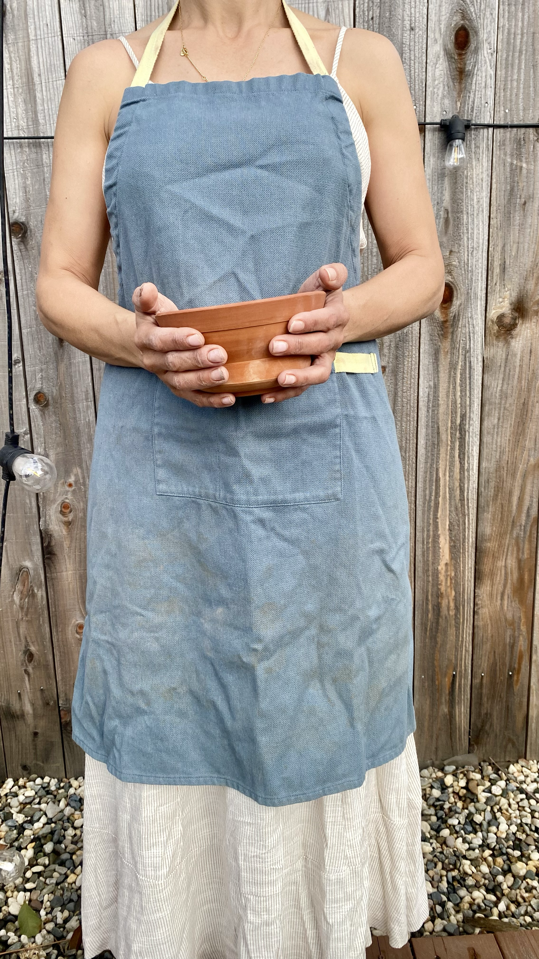 Person wearing a blue apron, holding a clay bowl, standing against a wooden fence with string lights.