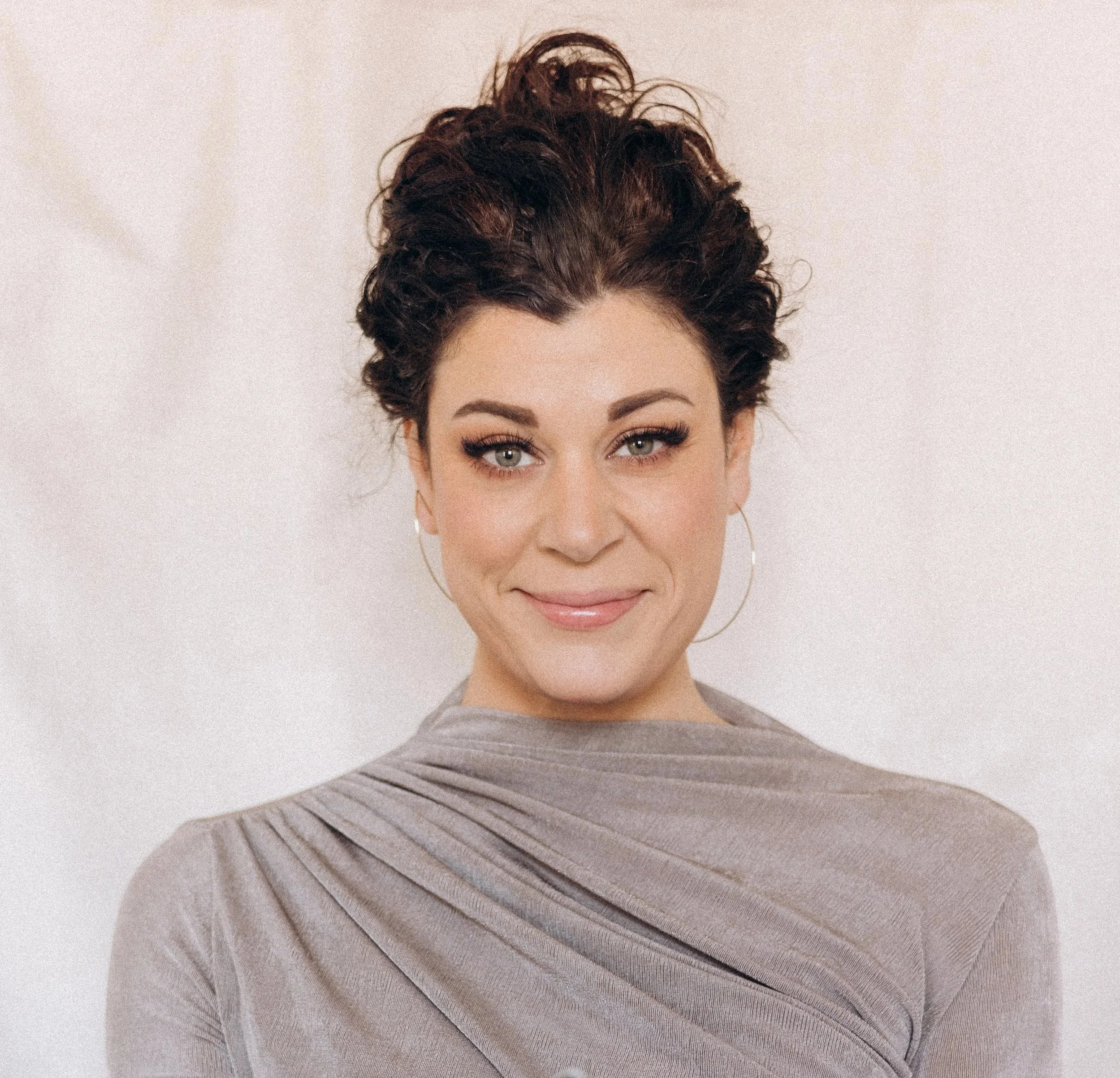 Portrait of a woman with curly dark brown hair, hoop earrings, and a light beige top, smiling softly against a neutral background.