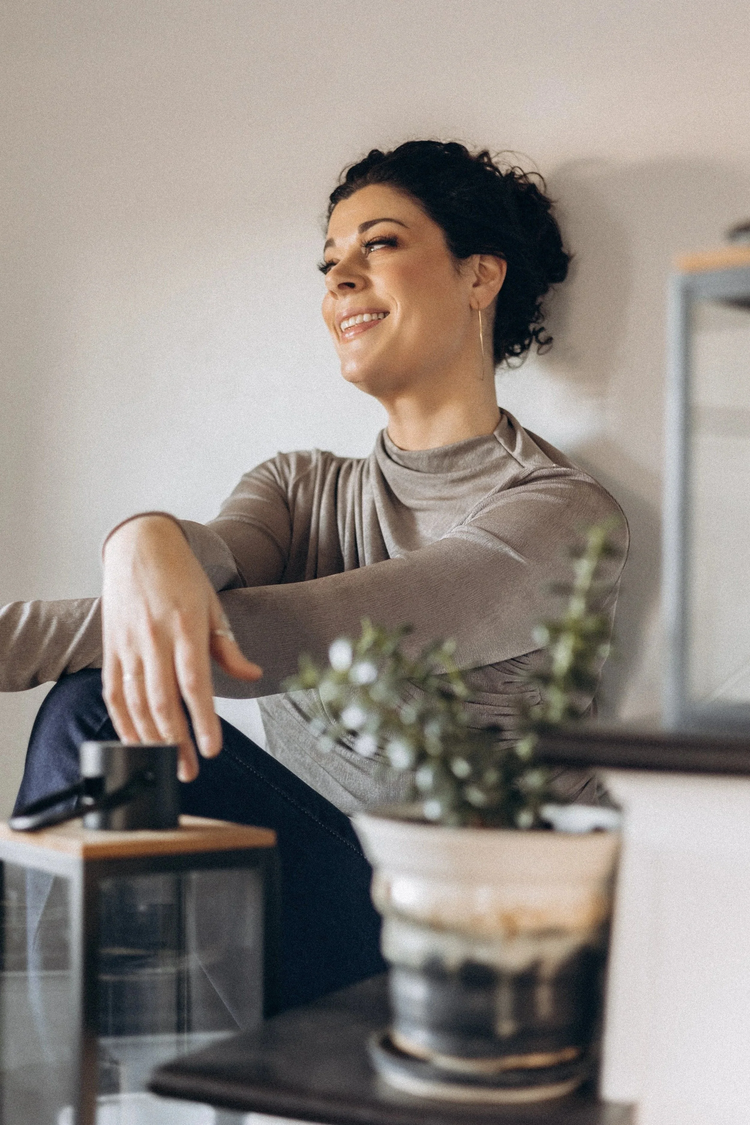 A woman with short curly dark hair smiling, sitting indoors against a white wall, wearing a beige silky top.