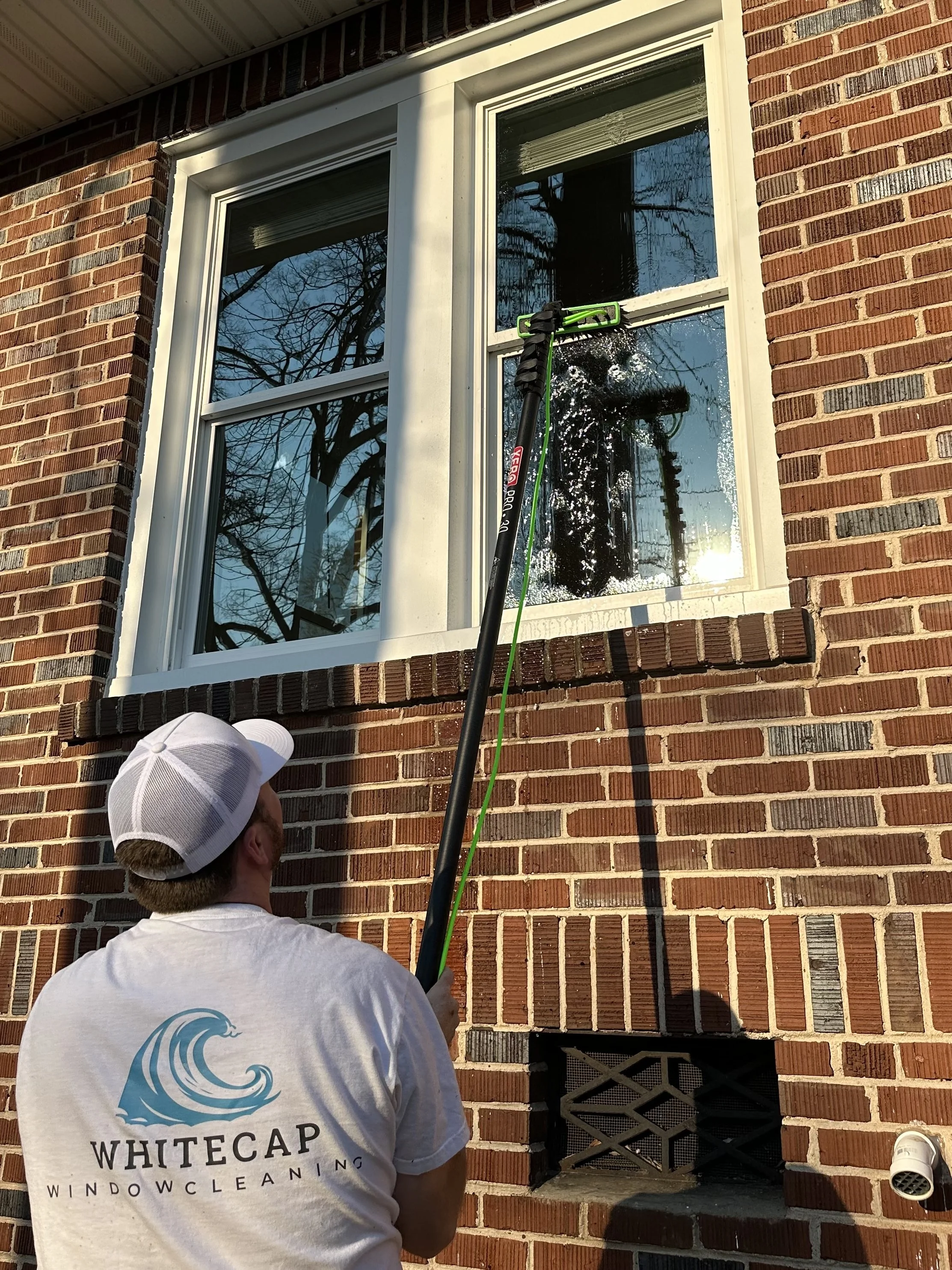 A person cleaning a window on a brick house using a long-handled squeegee, wearing a white cap and a white t-shirt with the logo 'Whitecap Window Cleaning.'
