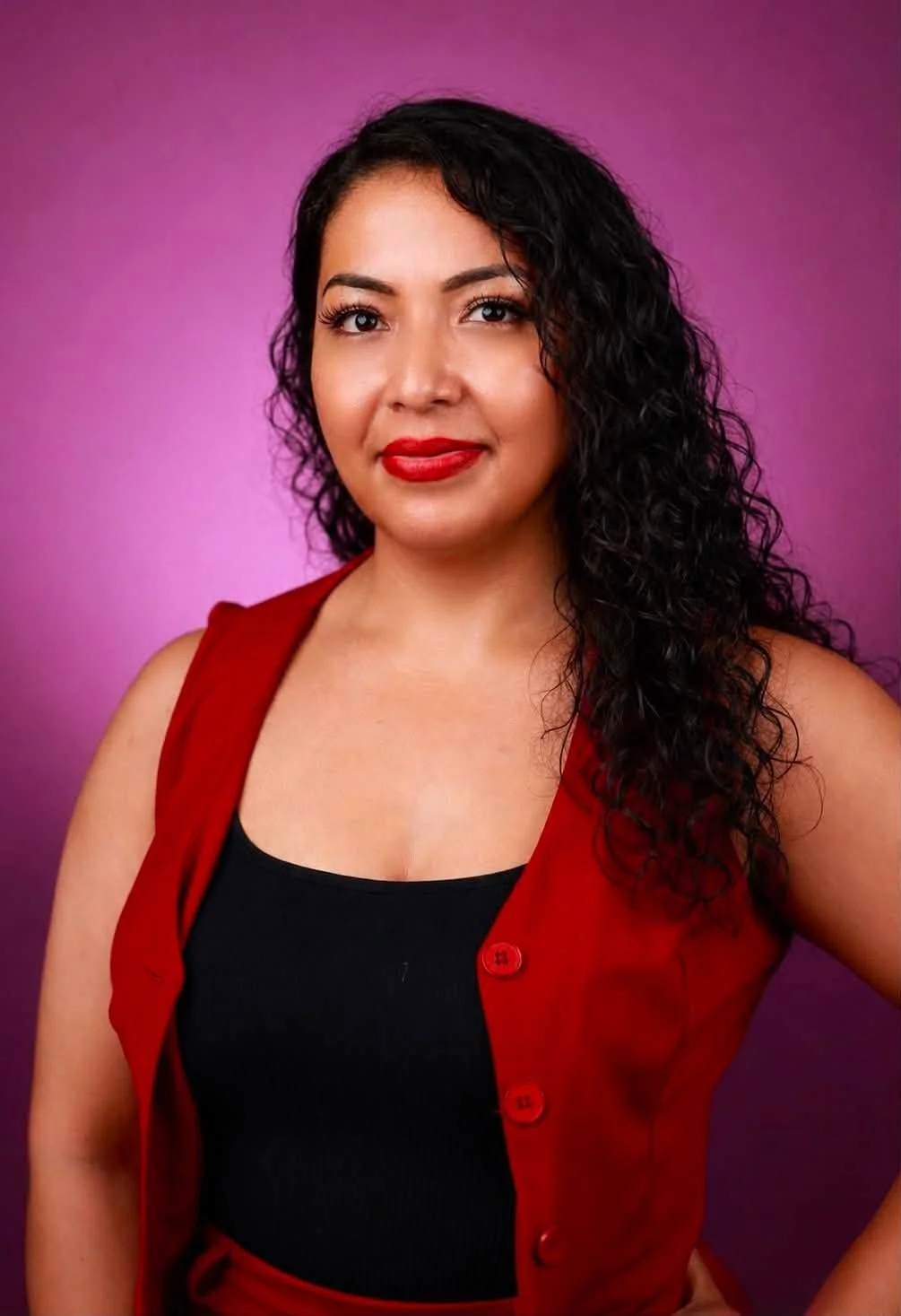 A woman with curly black hair, red lipstick, and wearing a black top with a red vest, standing against a purple background.