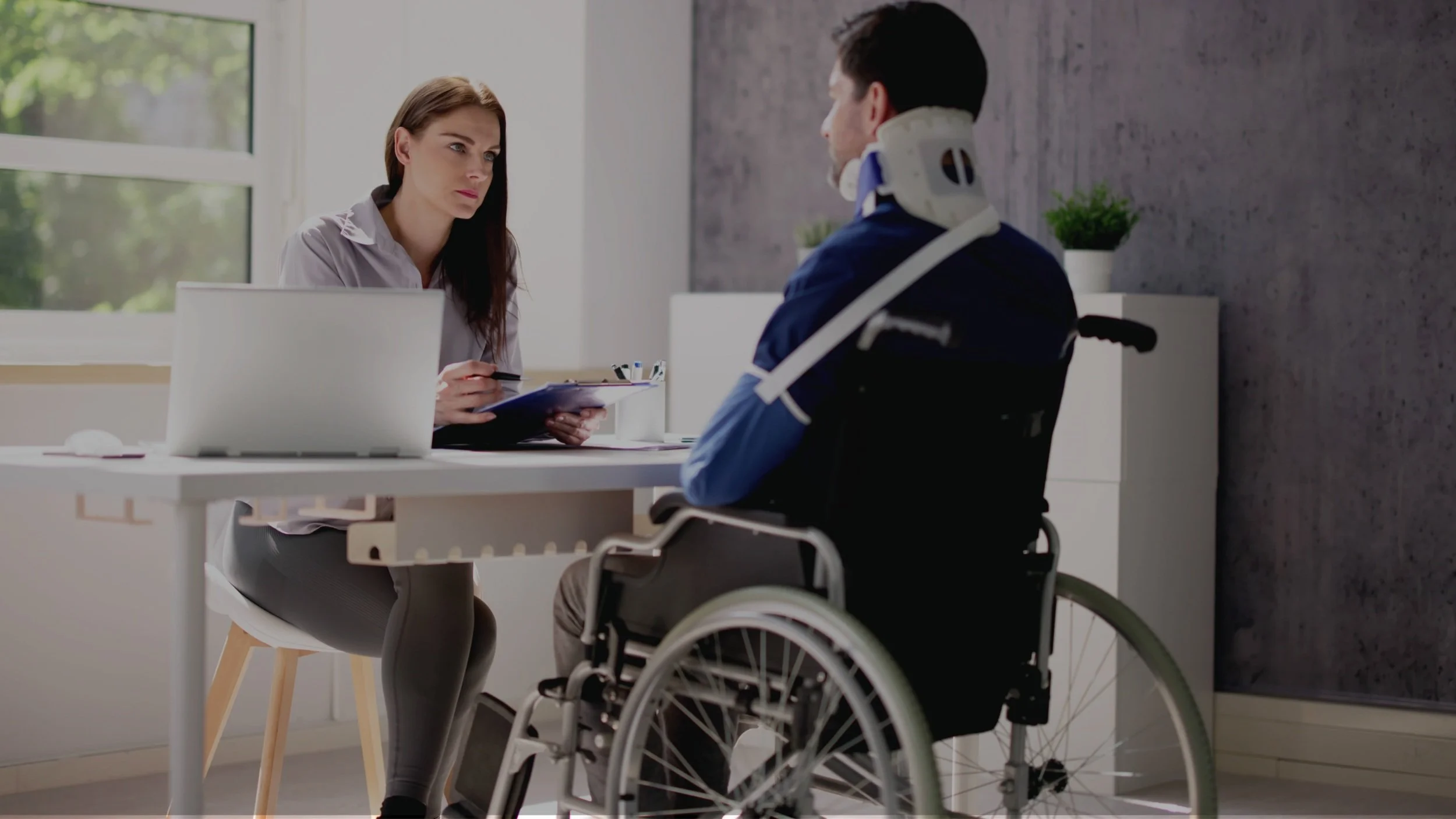 A woman sitting at a desk speaking with a man in a wheelchair who has a neck brace in a bright office with large windows and potted plants.