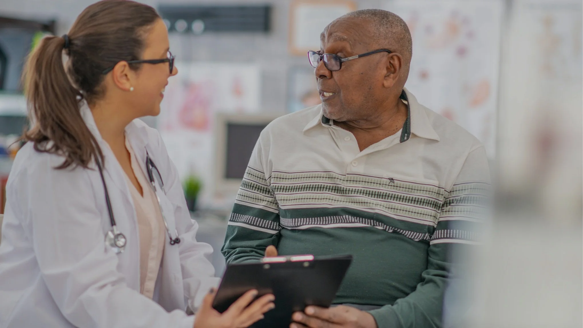 A female doctor and an elderly male patient having a conversation in a medical clinic, with the patient holding a tablet or clipboard.