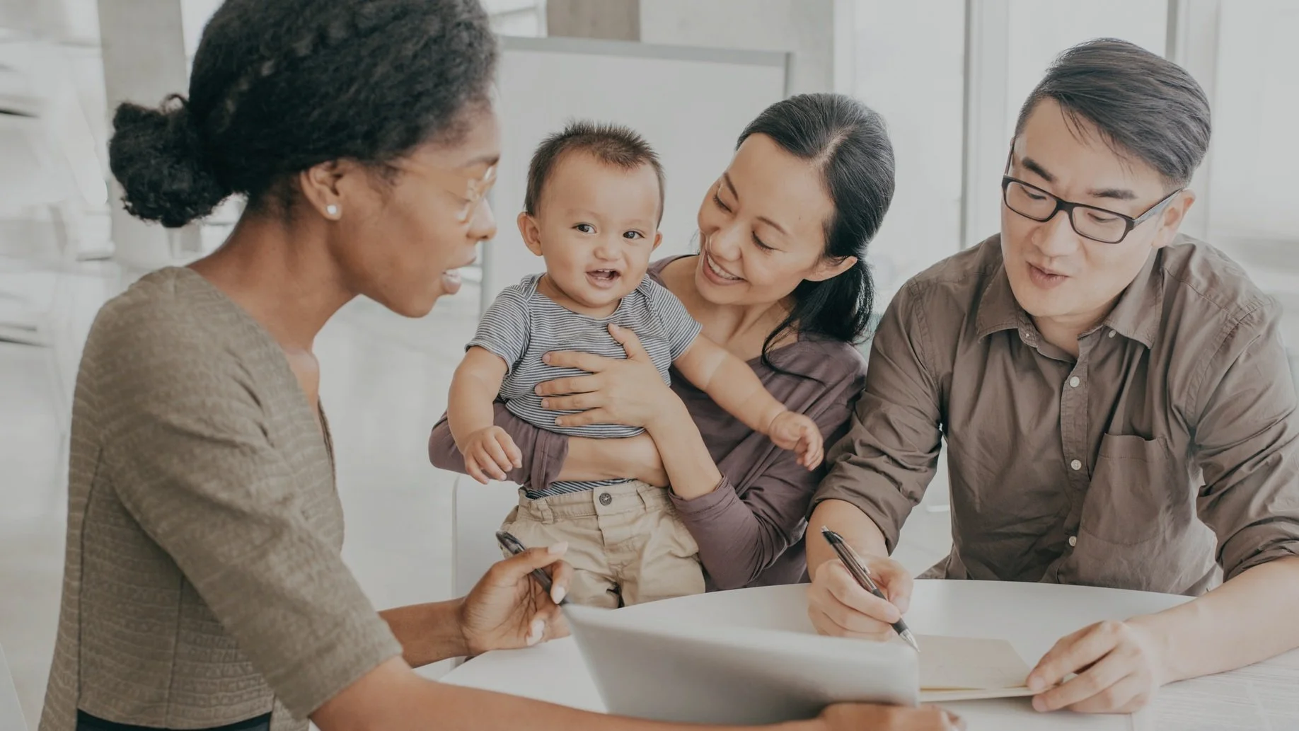 A diverse group of four adults and one child gathered around a table, engaging in a discussion or signing documents, with the child being held by one of the adults.