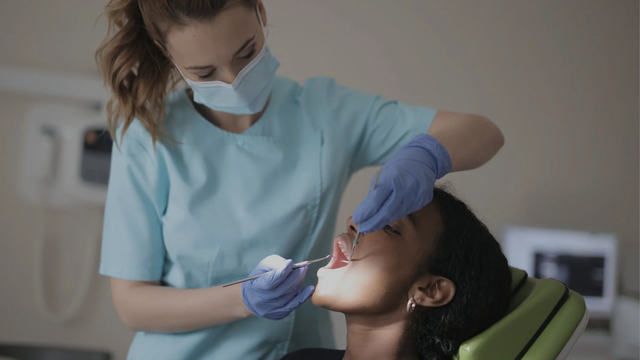 A dental professional in a face mask and gloves administers a dental procedure to a patient in a dental chair.