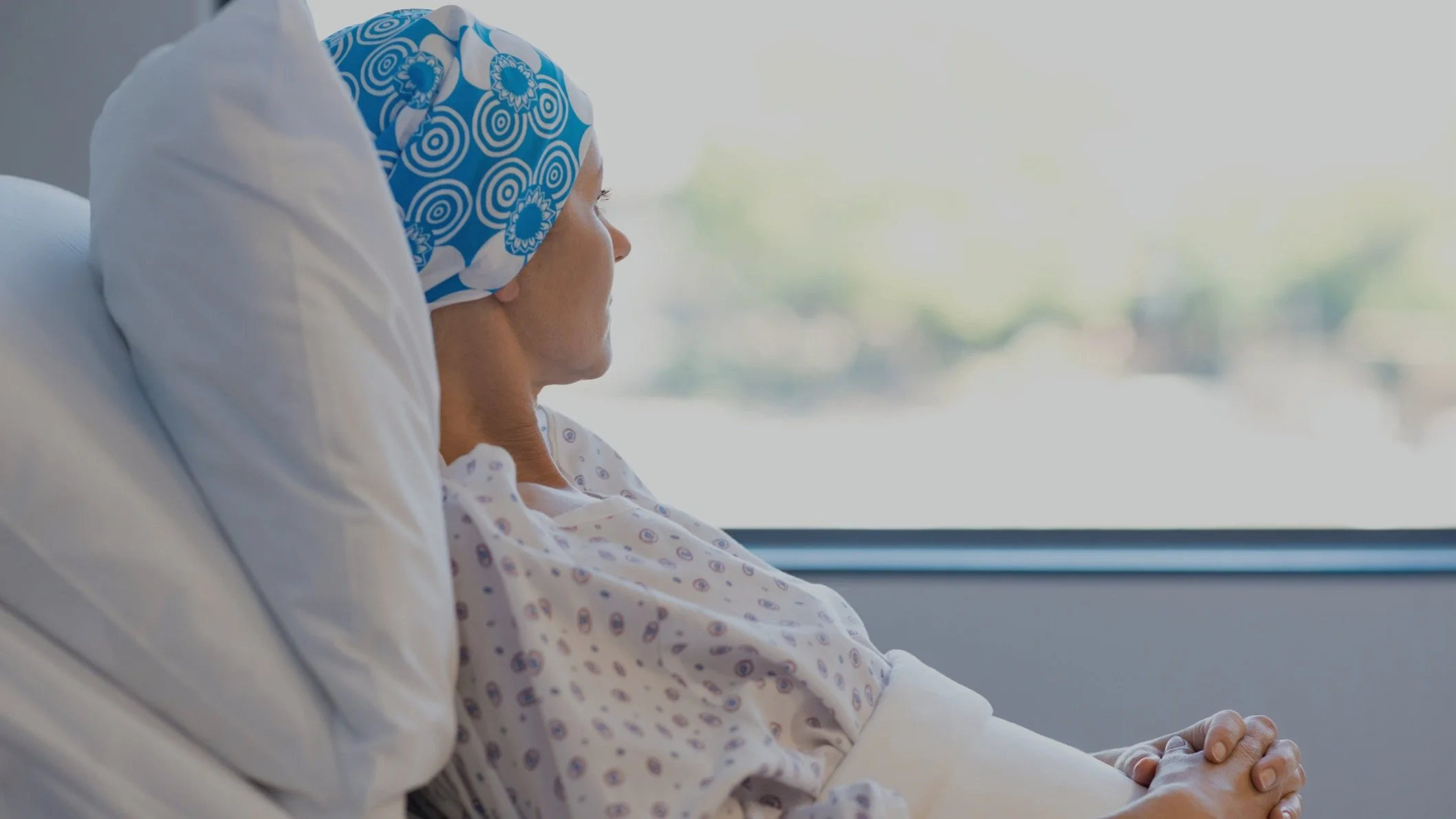 A woman in a hospital bed wearing a blue and white patterned surgical cap and gown, looking out the window.