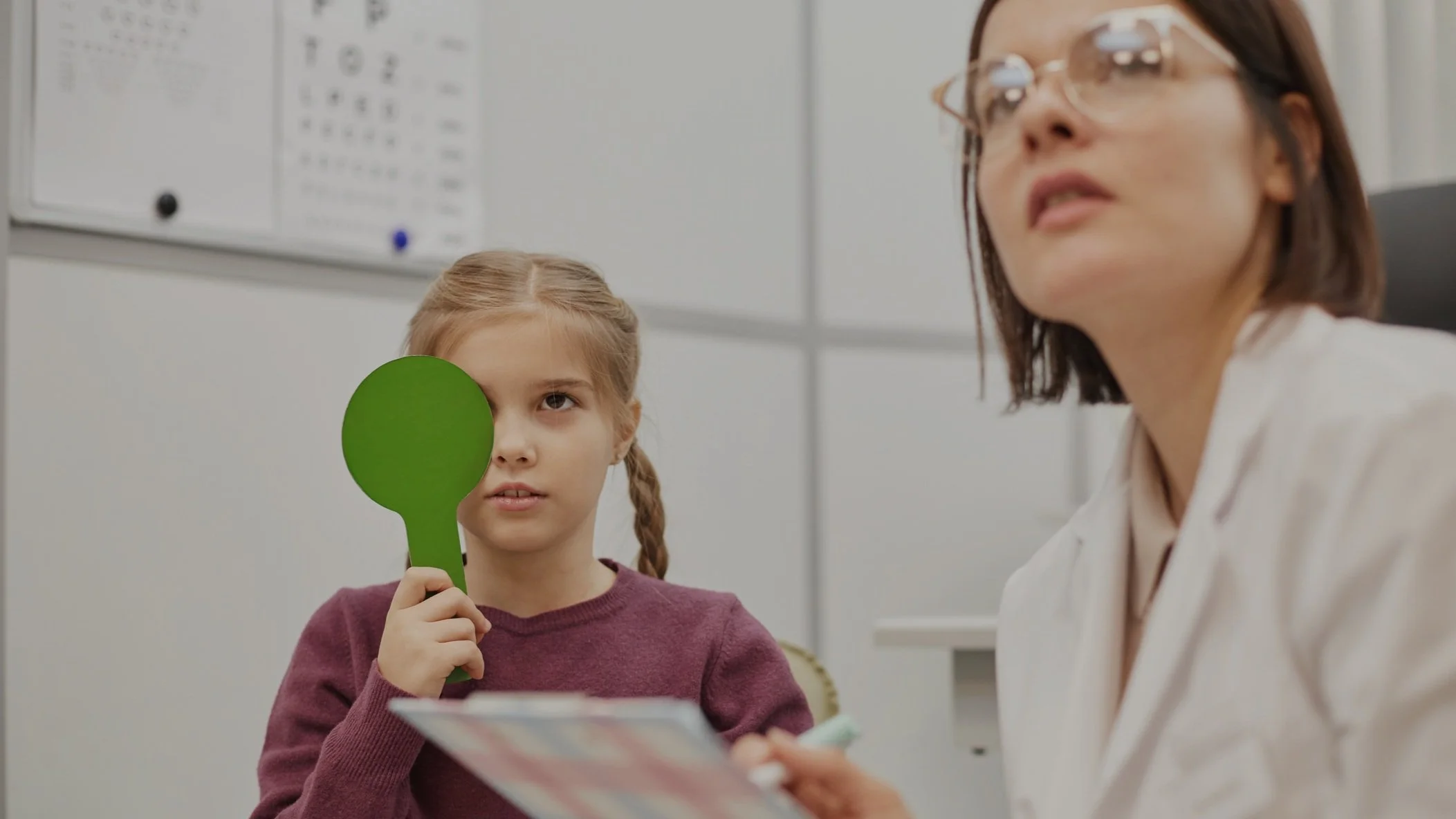 A young girl with braided hair holding a green paddle toy, looking at a woman, presumably a doctor or teacher, who is reading a book or paper.