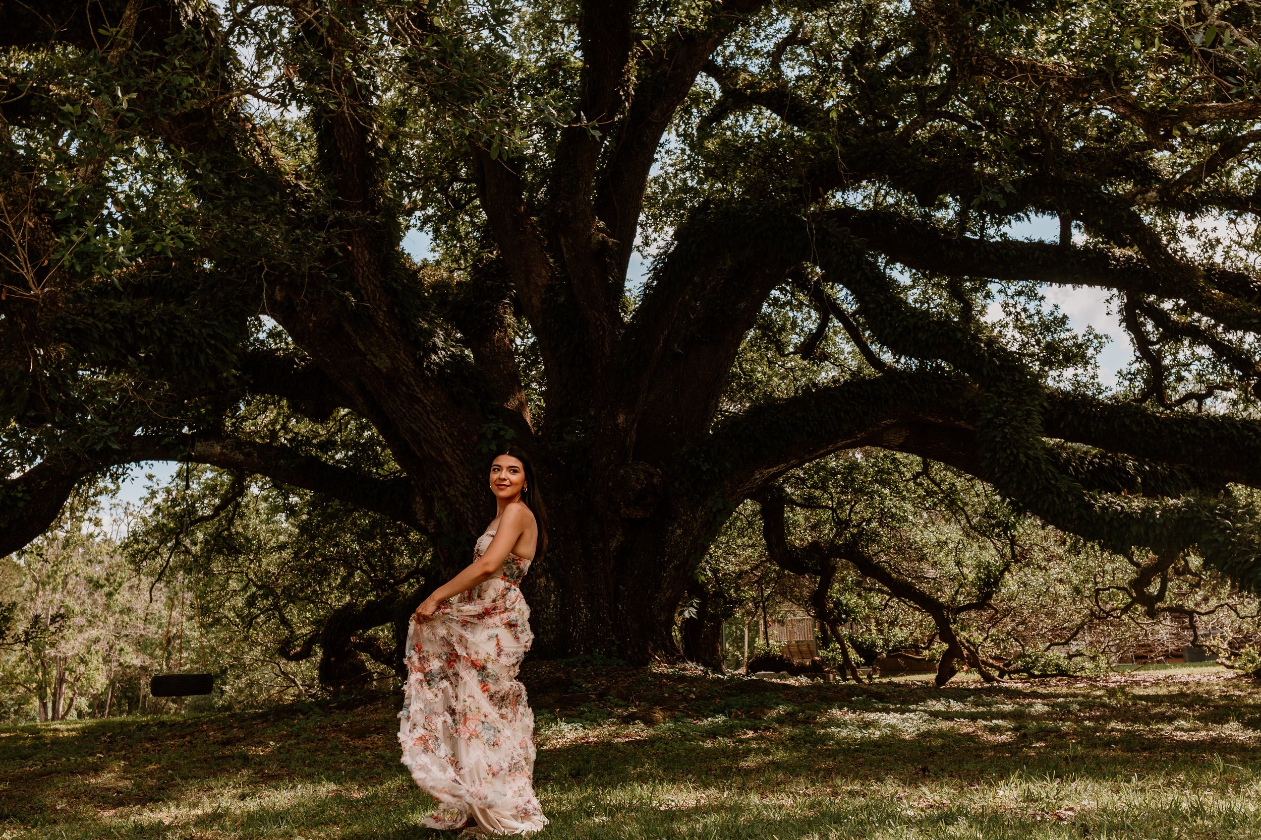 Woman in a flowing floral dress walking beneath a large oak tree during a creative portrait session in Central Mississippi. Photographed by Anna Klae.