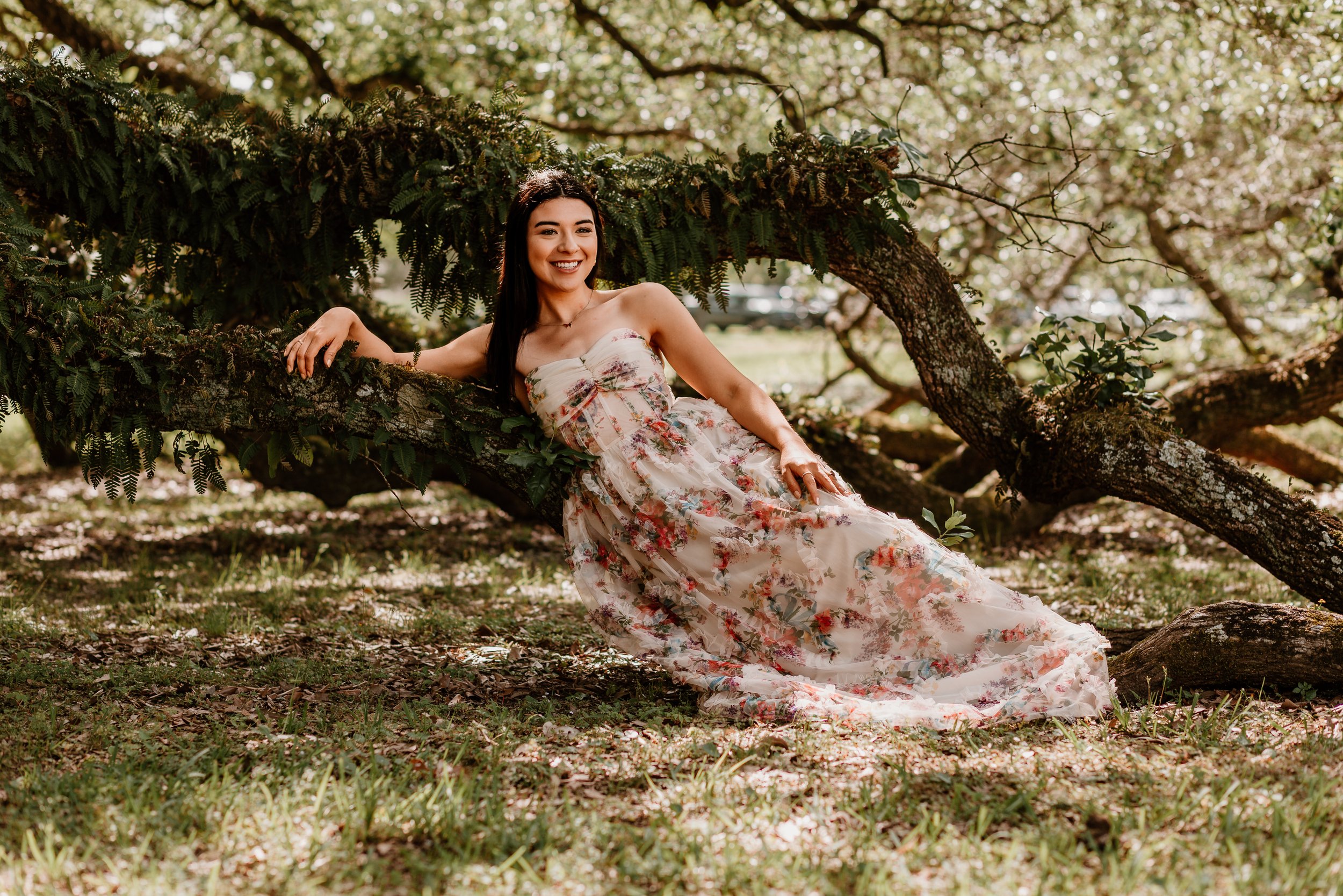 Woman in a flowing floral dress reclining on a moss-covered tree branch, smiling beneath a canopy of leaves in soft spring light.