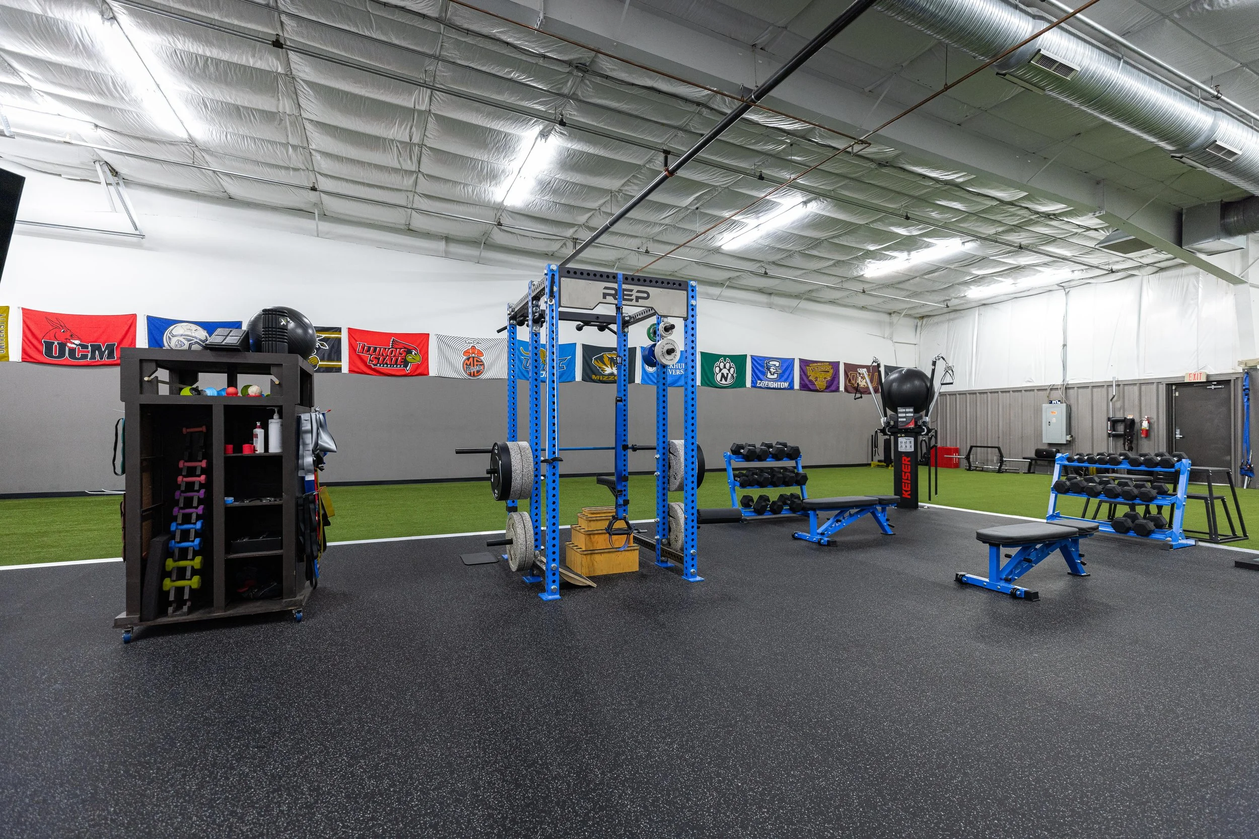 indoor gym with various weightlifting equipment including a blue squat rack, dumbbell racks, benches, and medicine balls, with sports team banners hanging on the wall.