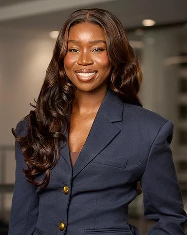 A smiling woman with long, wavy hair wearing a navy blazer with gold buttons, standing indoors.
