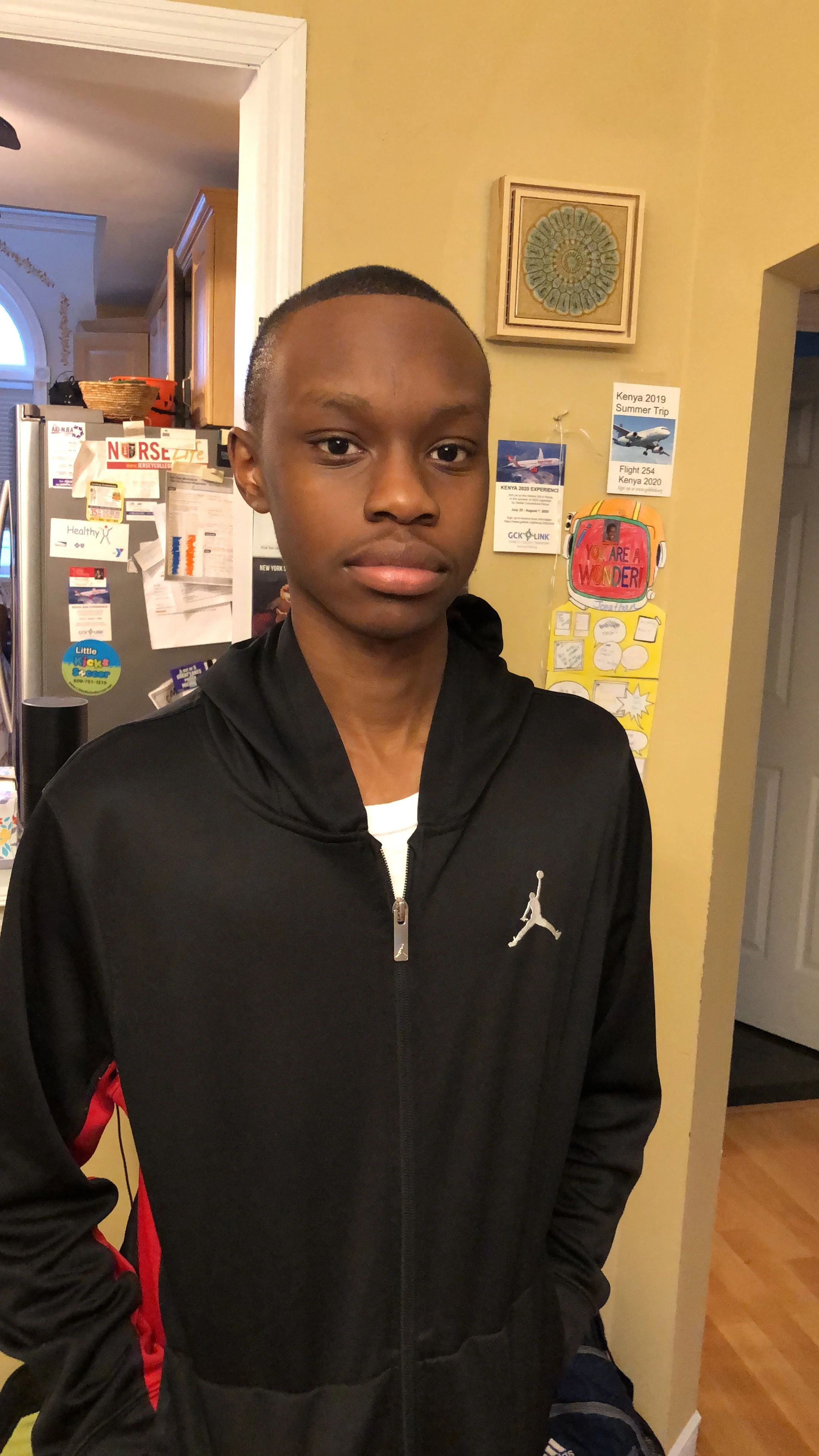 A young man standing indoors, wearing a black zip-up jacket with a white logo and a white shirt underneath. In the background, there is a yellow wall with various posters and decorations, including one with the text 'You Are a Wonder', and a refrigerator covered with notes and magnets.