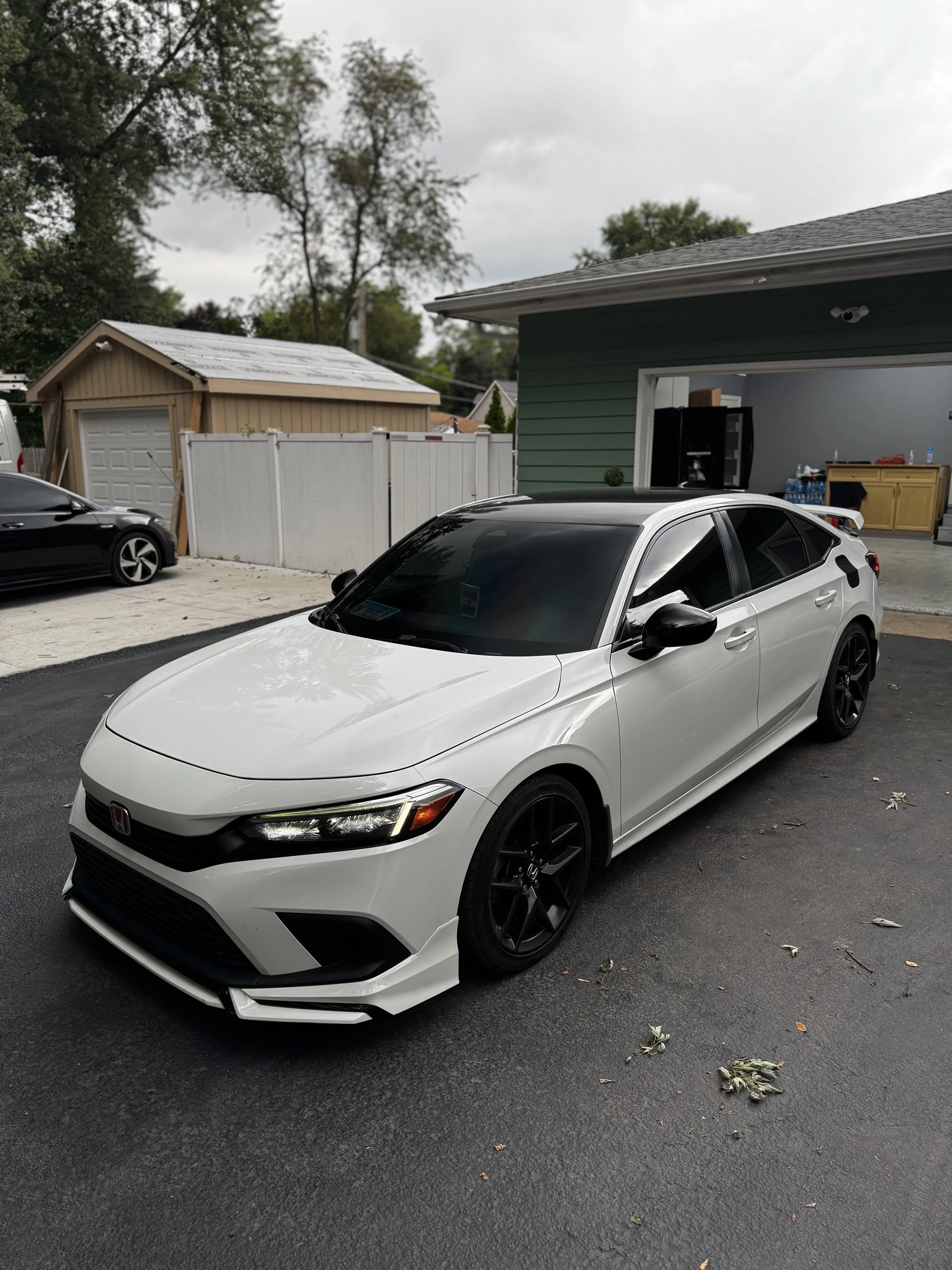 White Honda Civic parked on driveway in front of a house, with a black car to the side and a garage in the background.