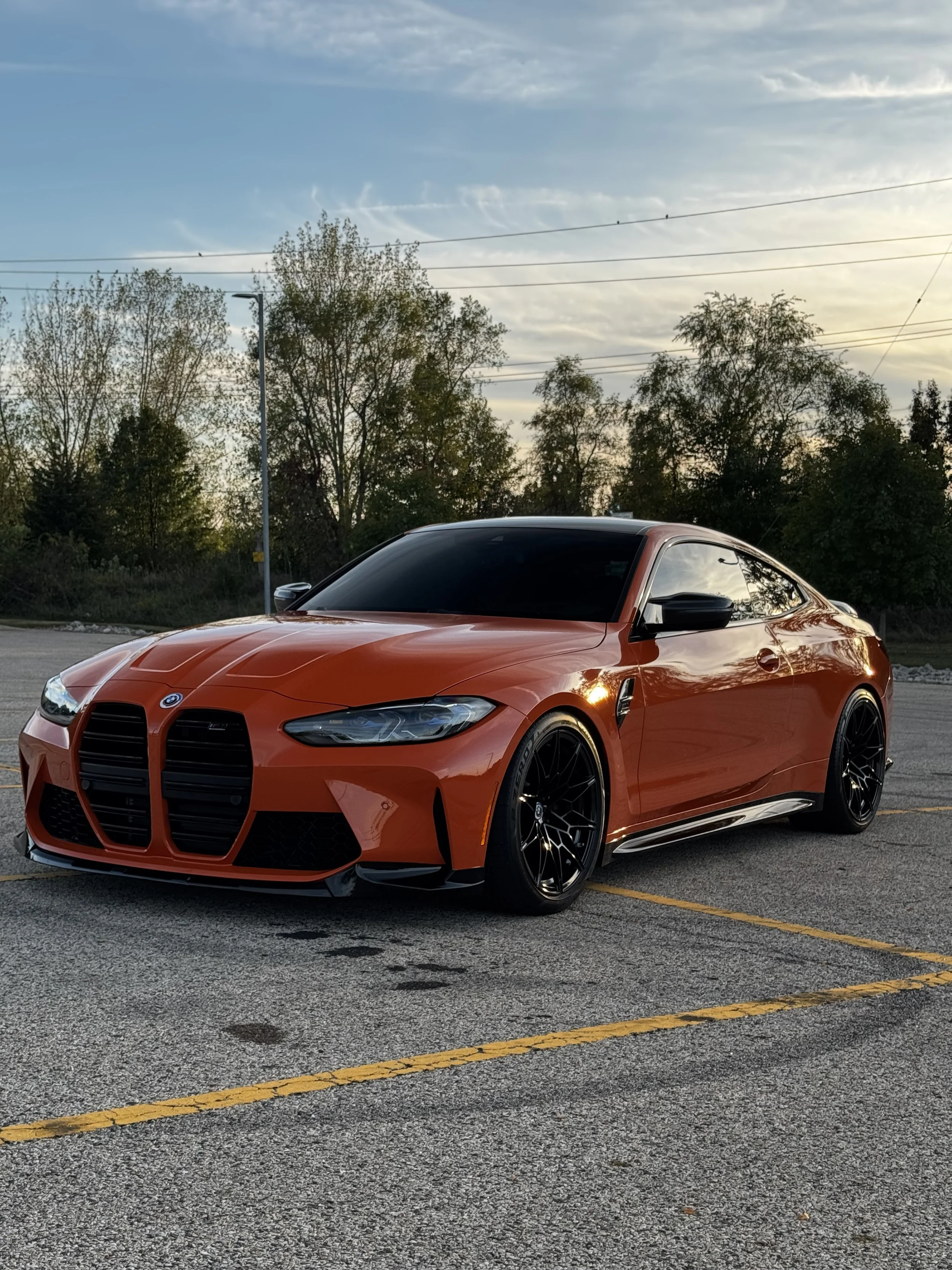 A bright orange sports car with black wheels parked in an empty parking lot, with trees and a partly cloudy sky in the background during sunset.