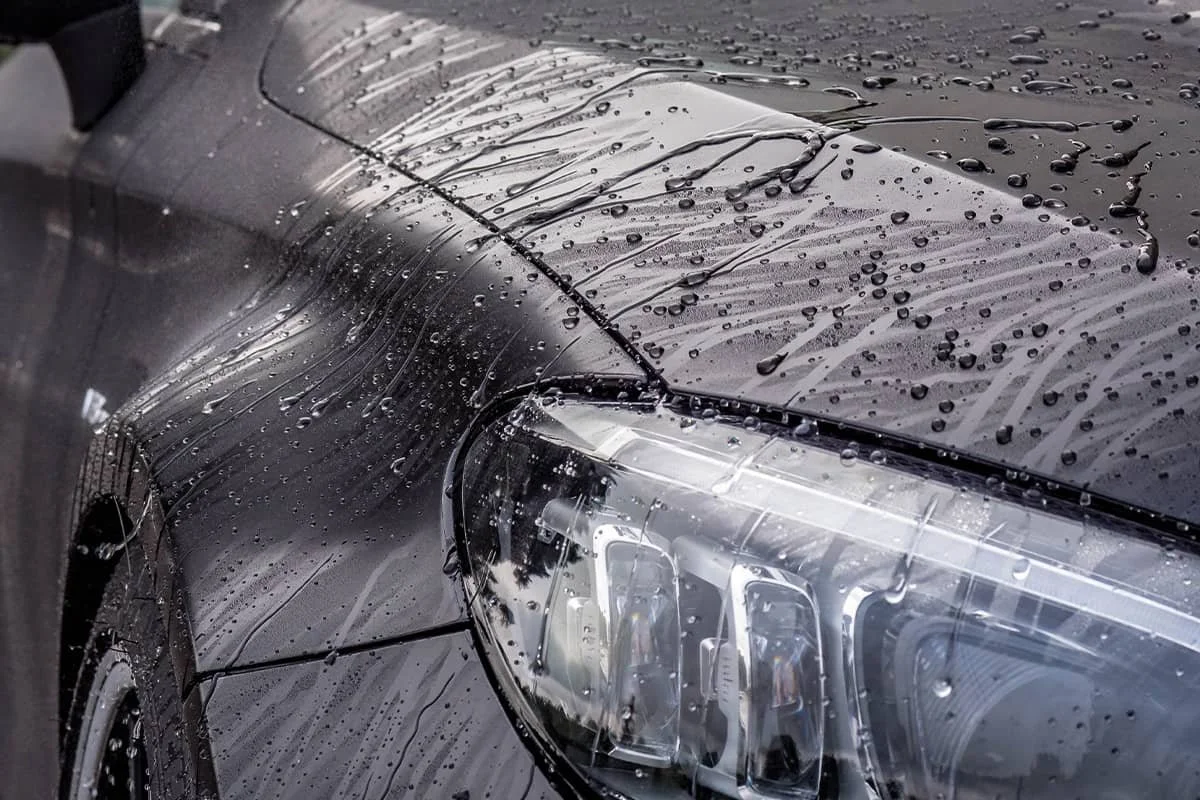 Close-up of a black car's front with water droplets and streaks on the hood and headlight.
