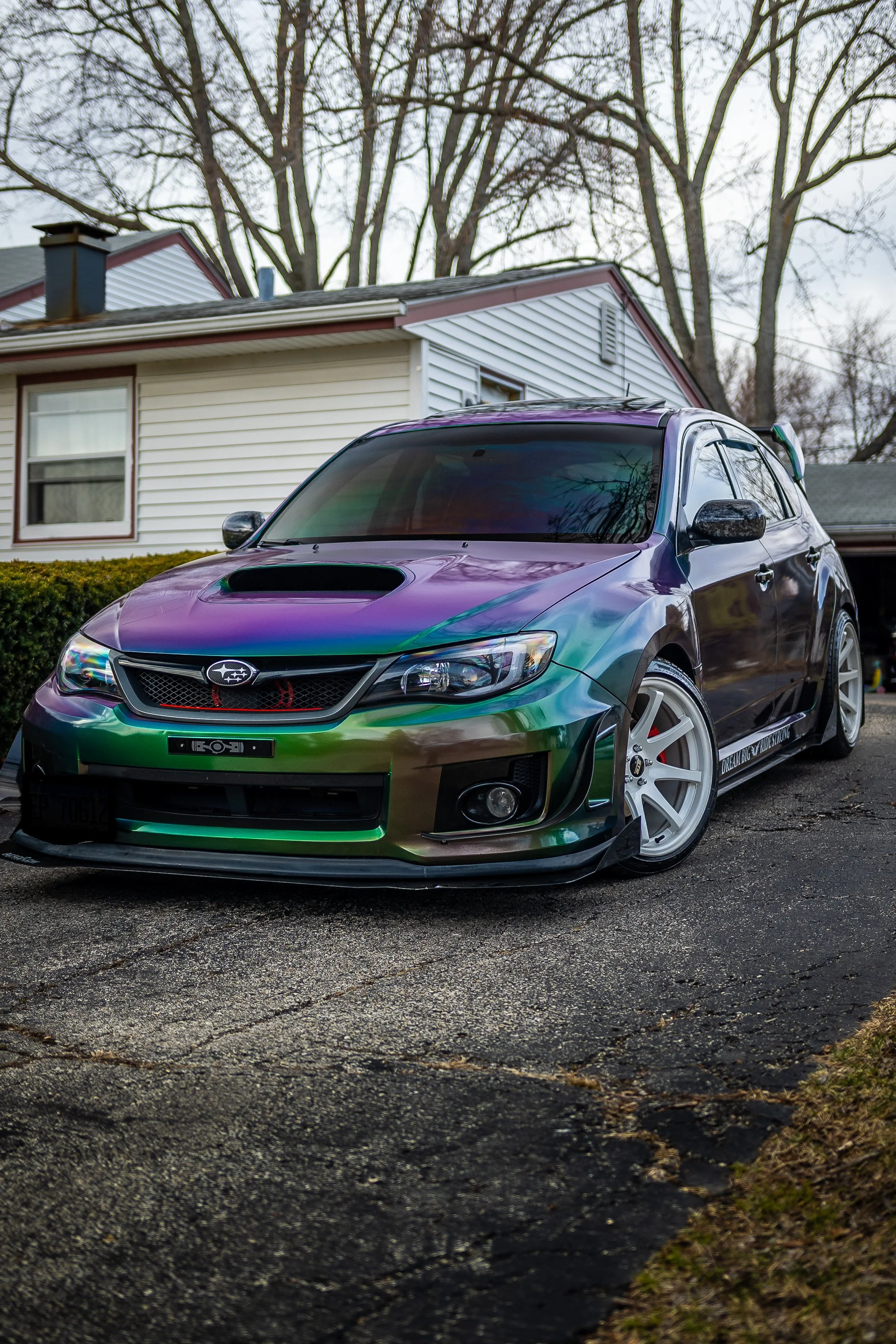 A colorful modified Subaru car parked on a driveway in front of a house, with leafless trees in the background.