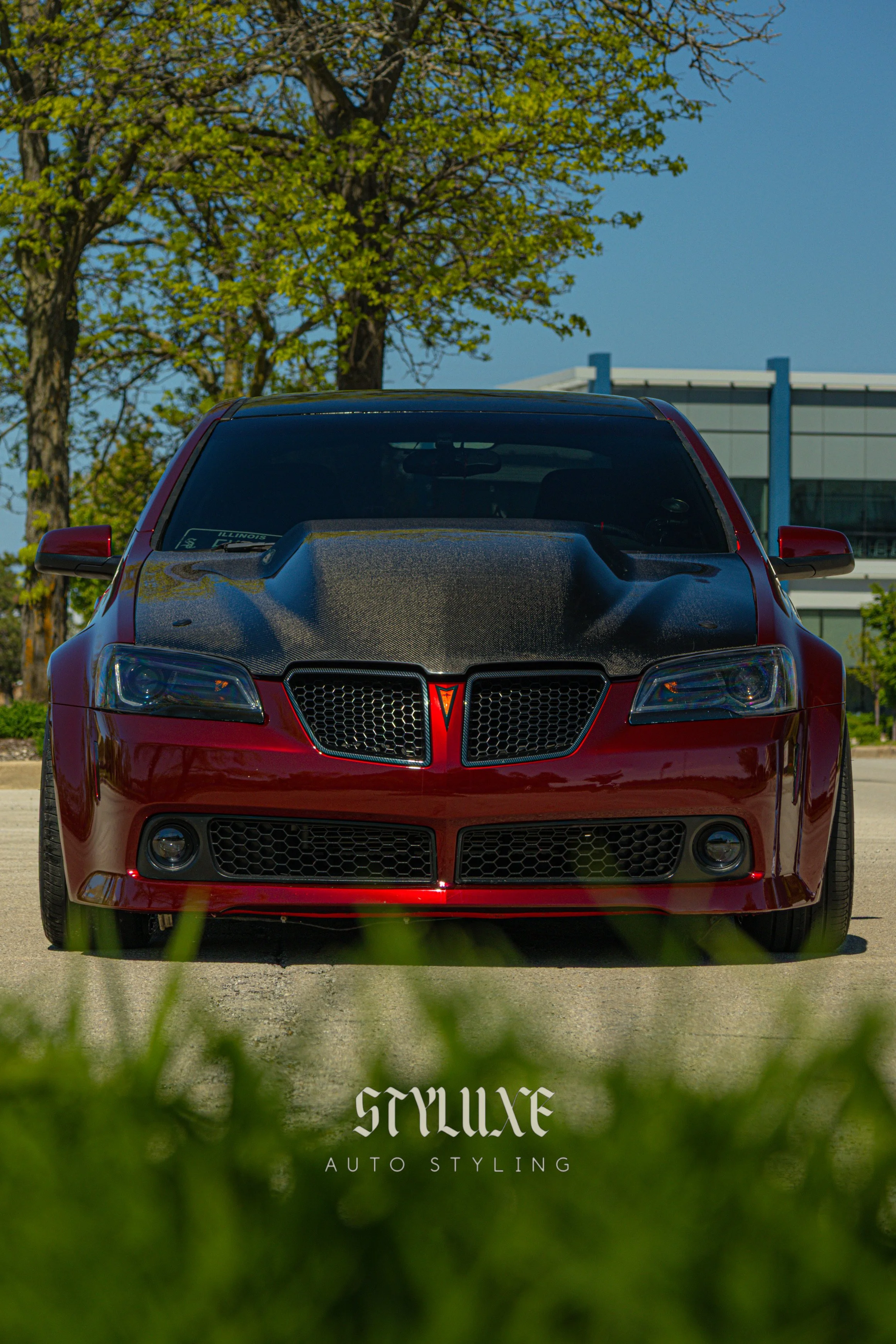 Red sports car with a black hood parked outdoors with trees and a building in the background, and the text 'StyLux Auto Styling' at the bottom.