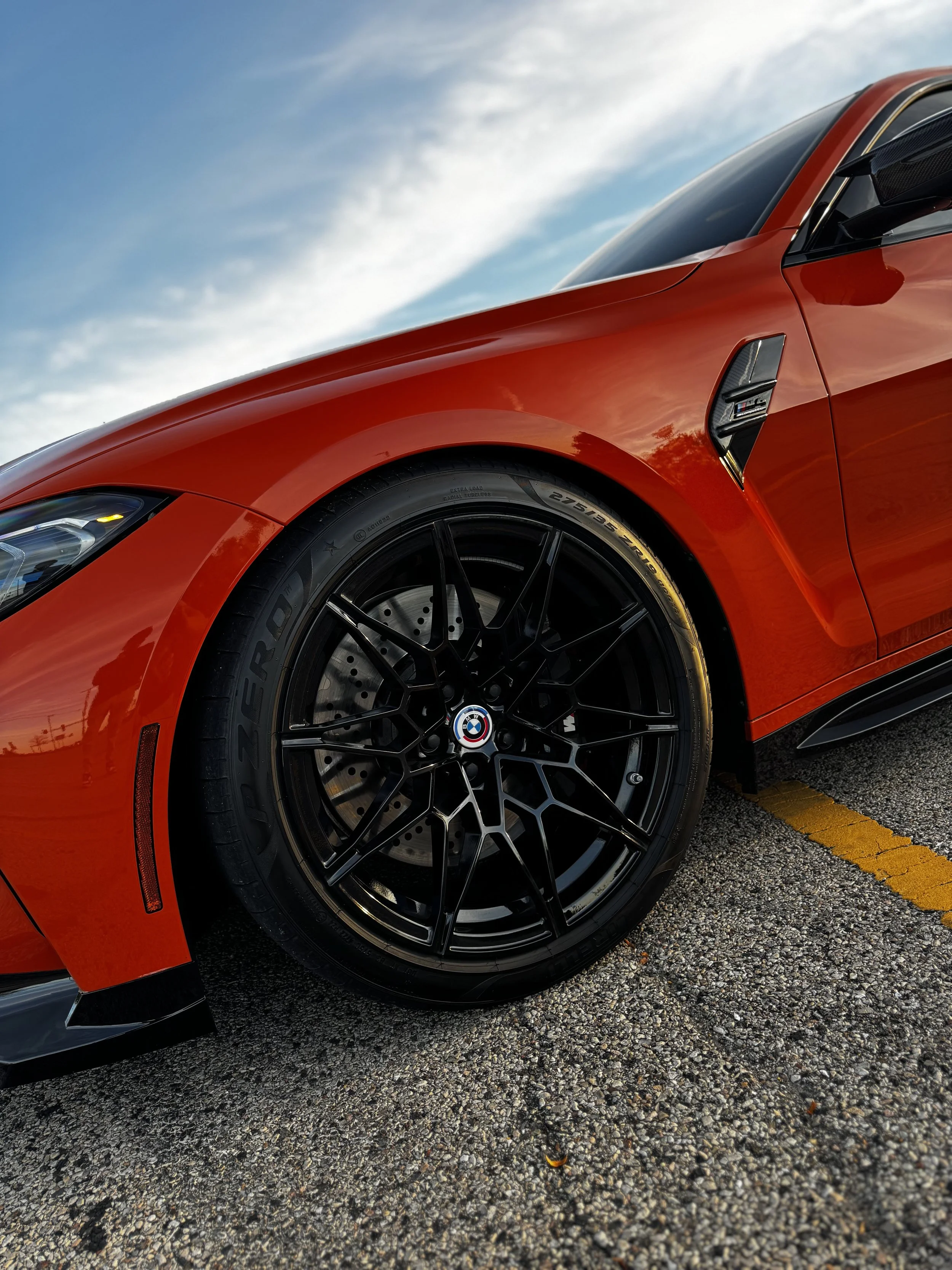 Close-up of front part of red BMW sports car with black wheels, parked on asphalt with yellow parking line, under a partly cloudy sky.