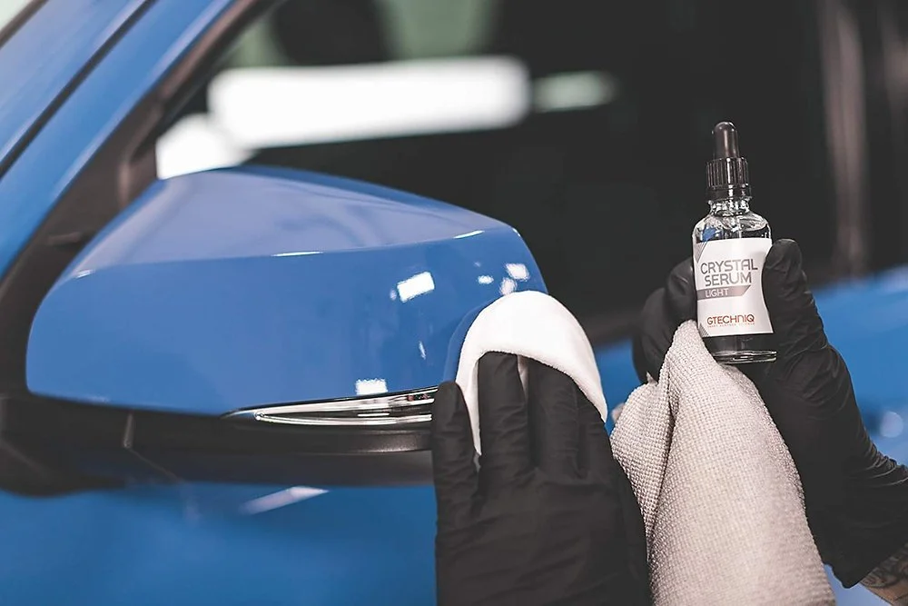 Person wearing black gloves cleaning a blue car's side mirror with a white cloth and a bottle of crystal serum inside a dropper bottle.