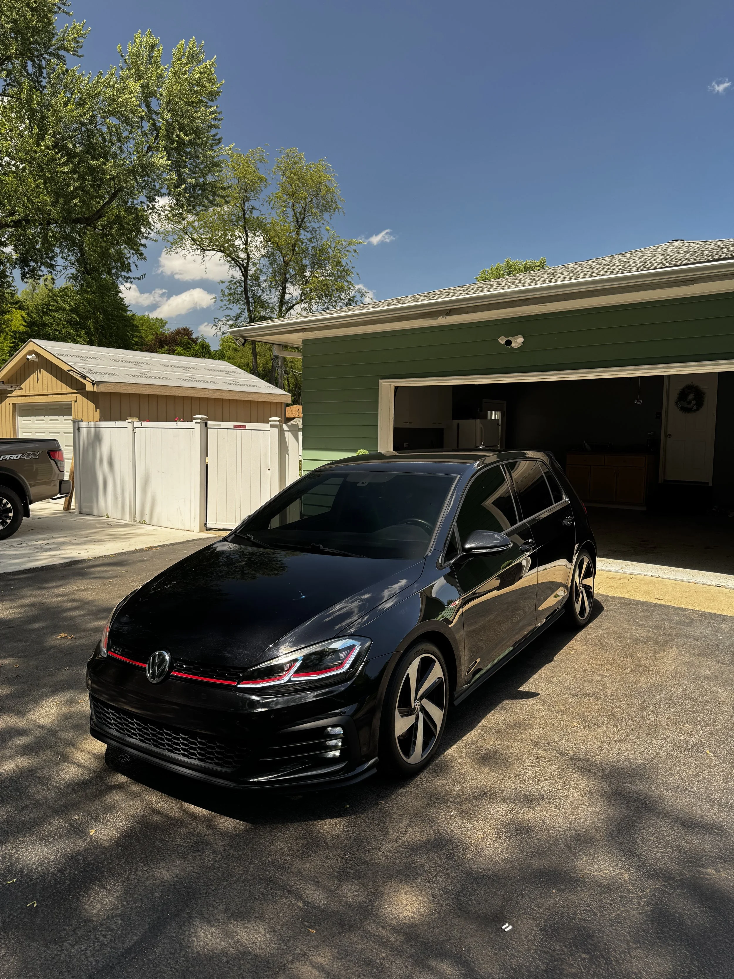 A black Volkswagen Golf parked in front of a green garage on a sunny day. There are trees and a small shed in the background, and the sky is blue with some clouds.