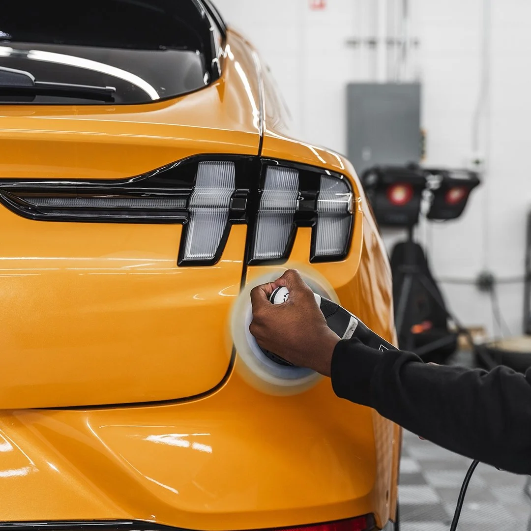 A person polishing the rear of a yellow sports car with a power buffer in a garage.
