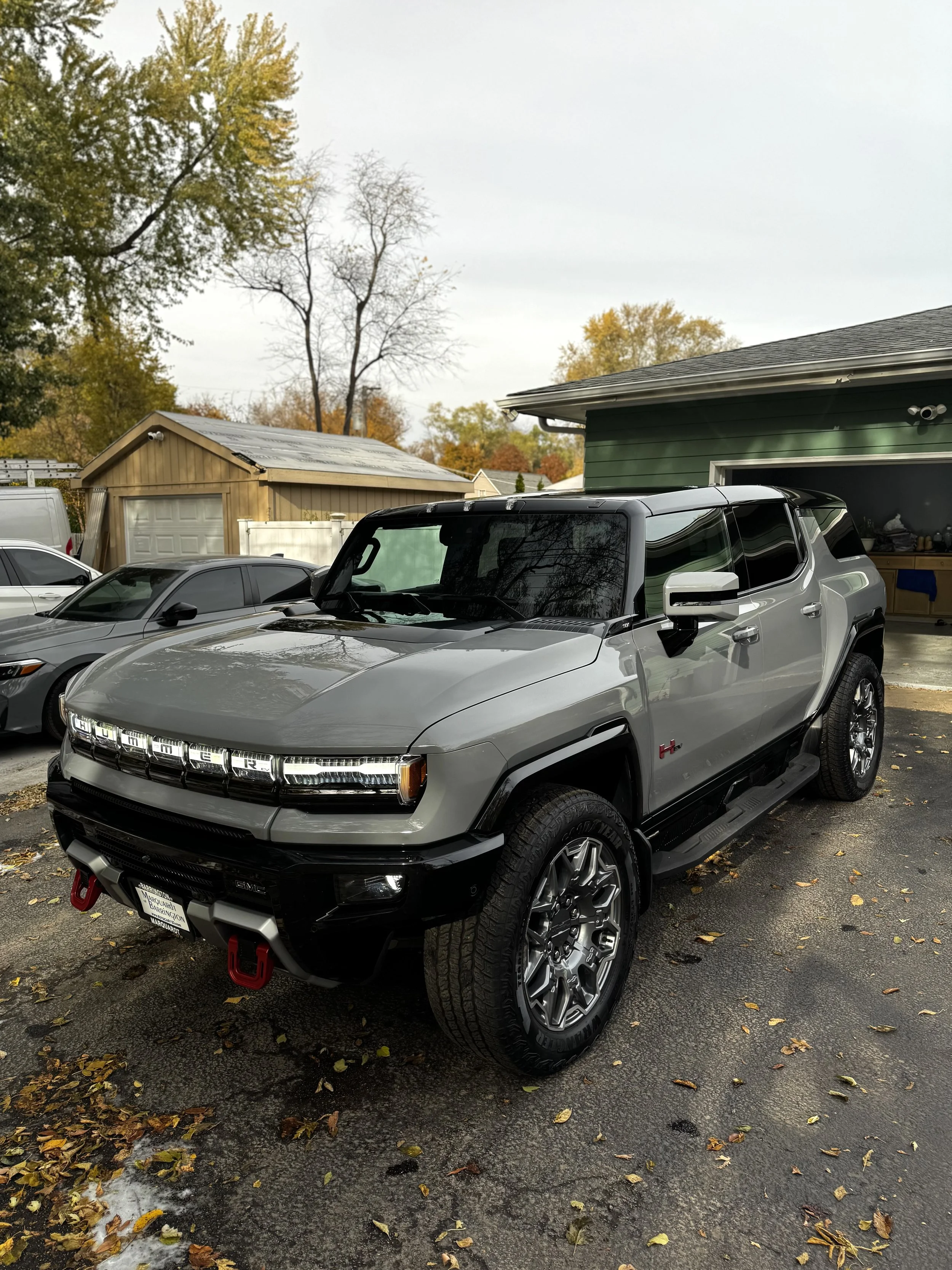 A silver and black GMC Hummer EV electric pickup truck parked in a driveway with fallen leaves, houses, trees with autumn foliage, and a storage shed in the background.
