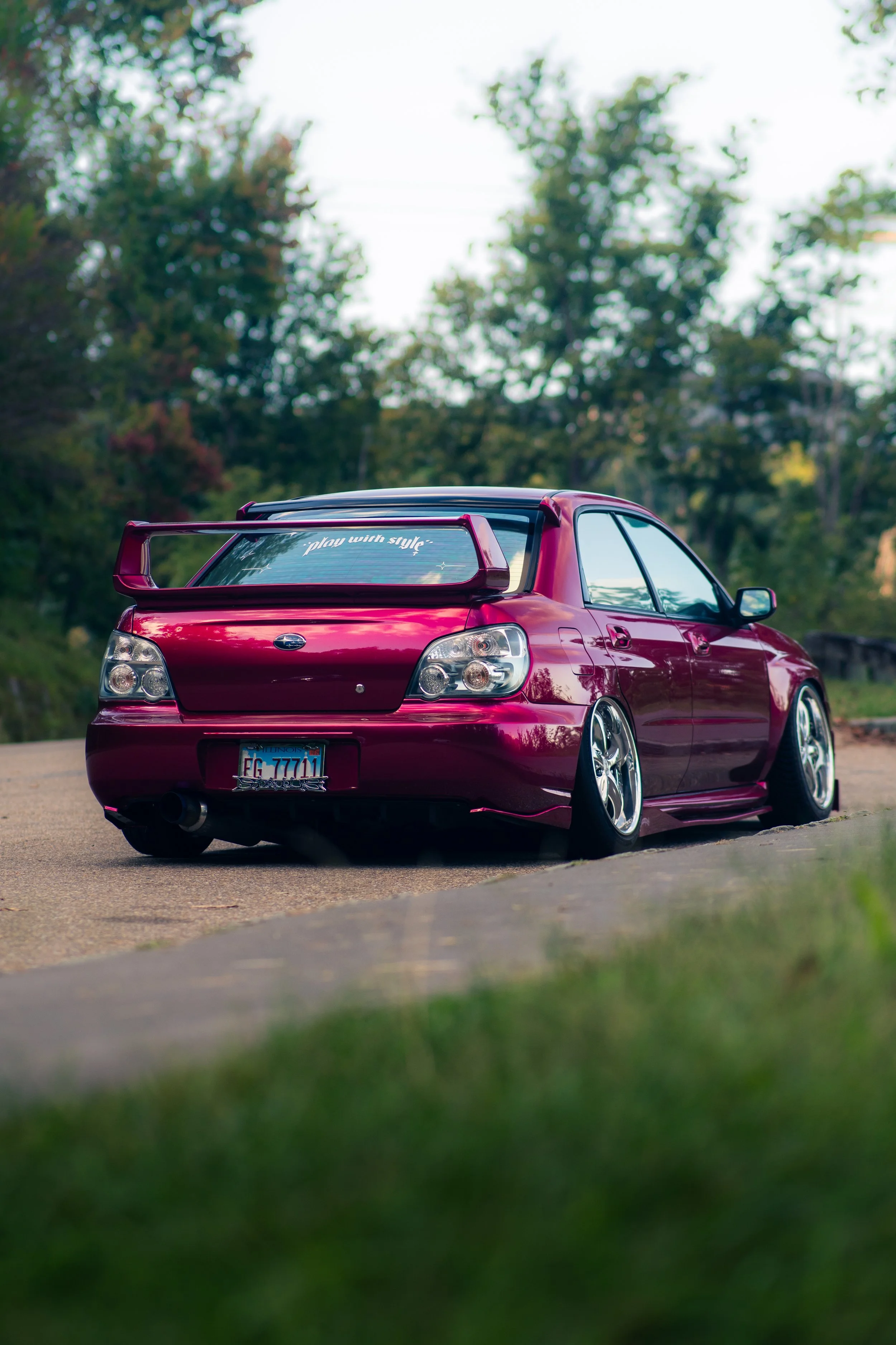 A shiny, modified red sedan with a large rear spoiler parked on a paved suburban street with green trees in the background.
