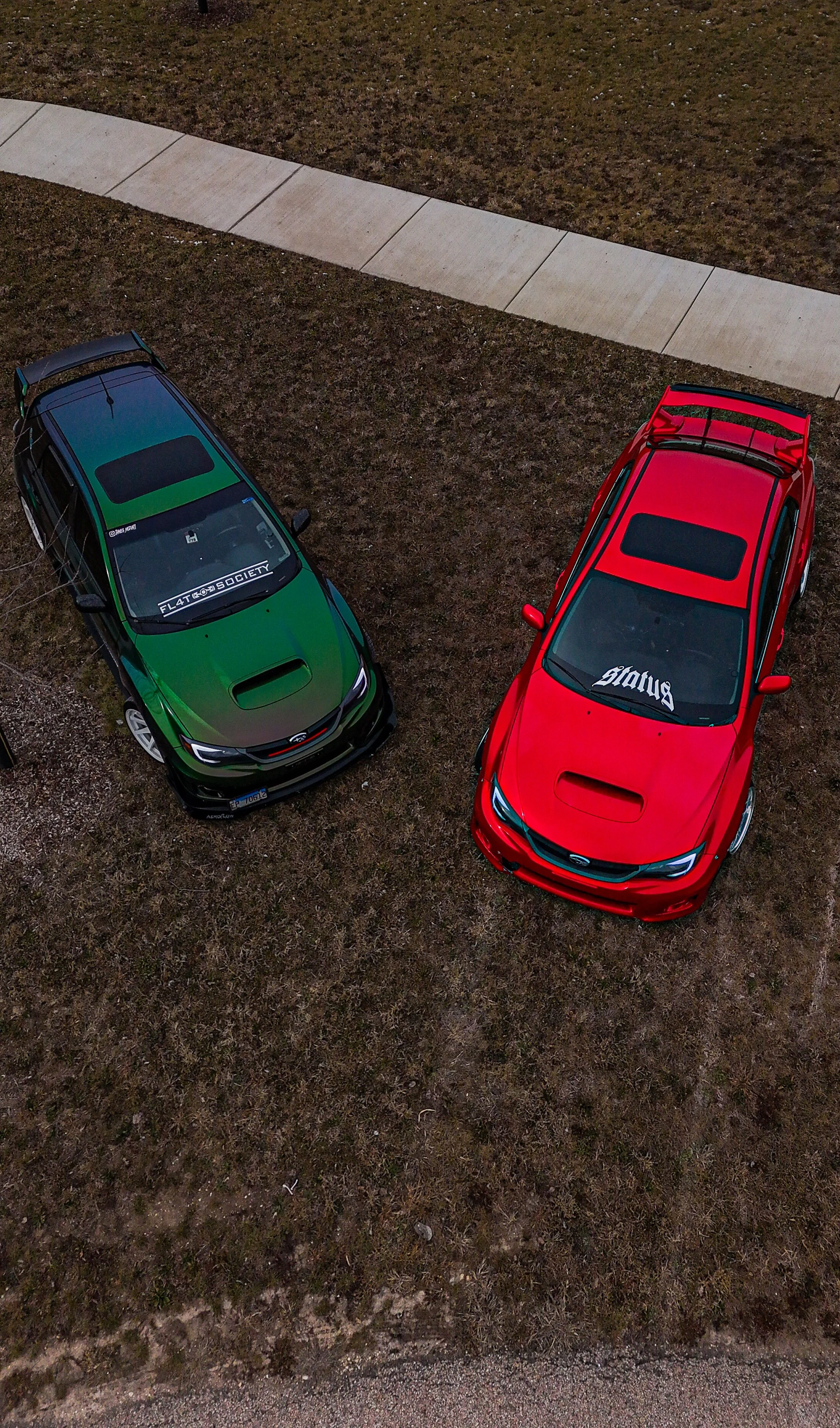 Aerial view of two cars, one green and black and one red, parked on a grassy area with a concrete sidewalk in the background.