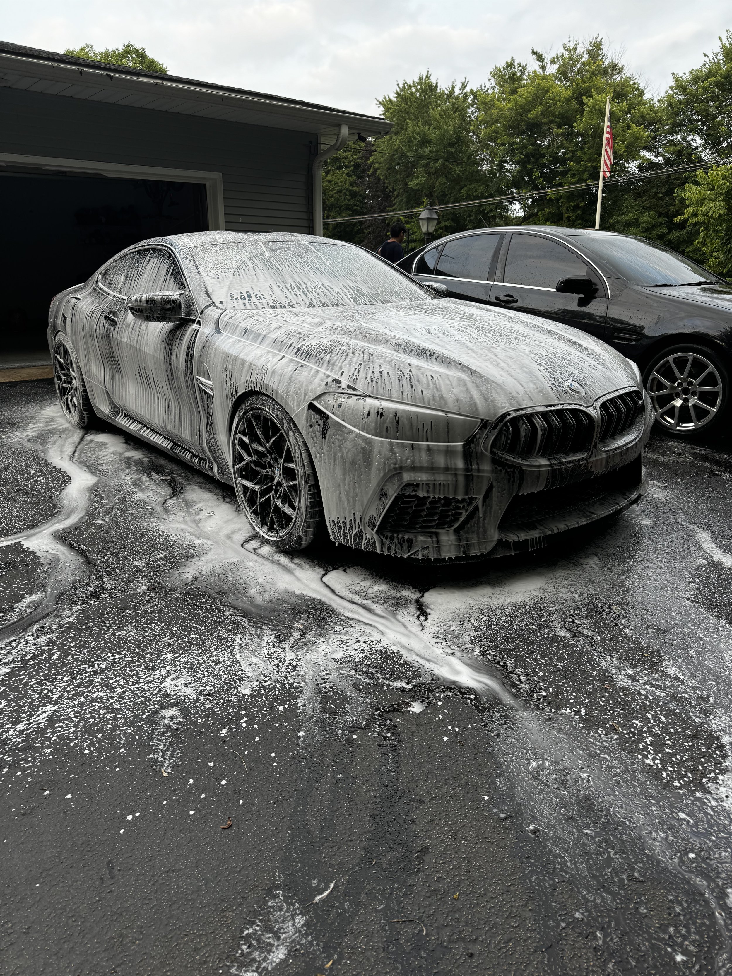A black sports car being washed with soap and foam on its surface in a driveway, with another black car parked beside it and trees in the background.