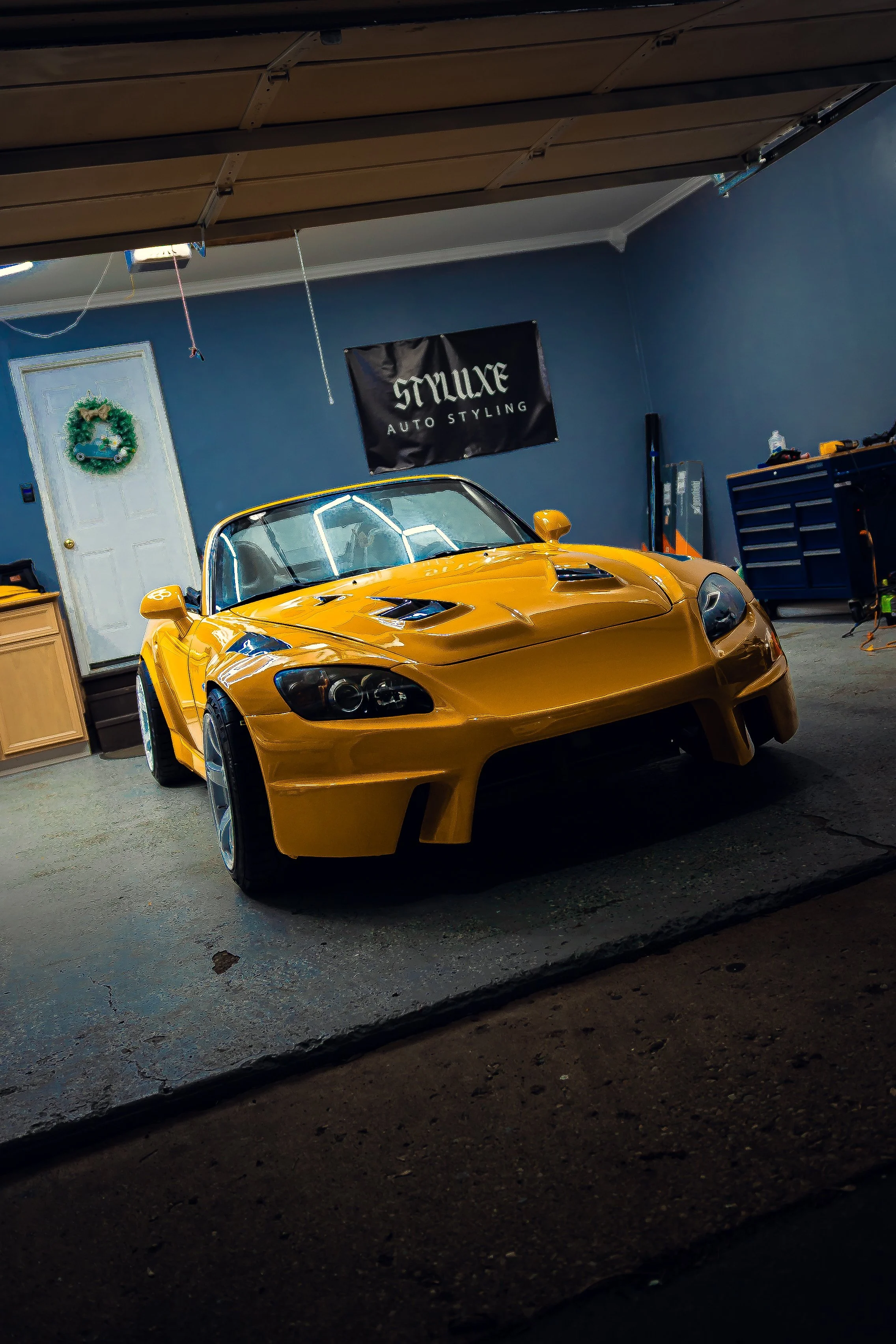 A yellow sports car inside a garage with a hanging sign that reads 'STYLEUX AUTO STYLING' behind it.
