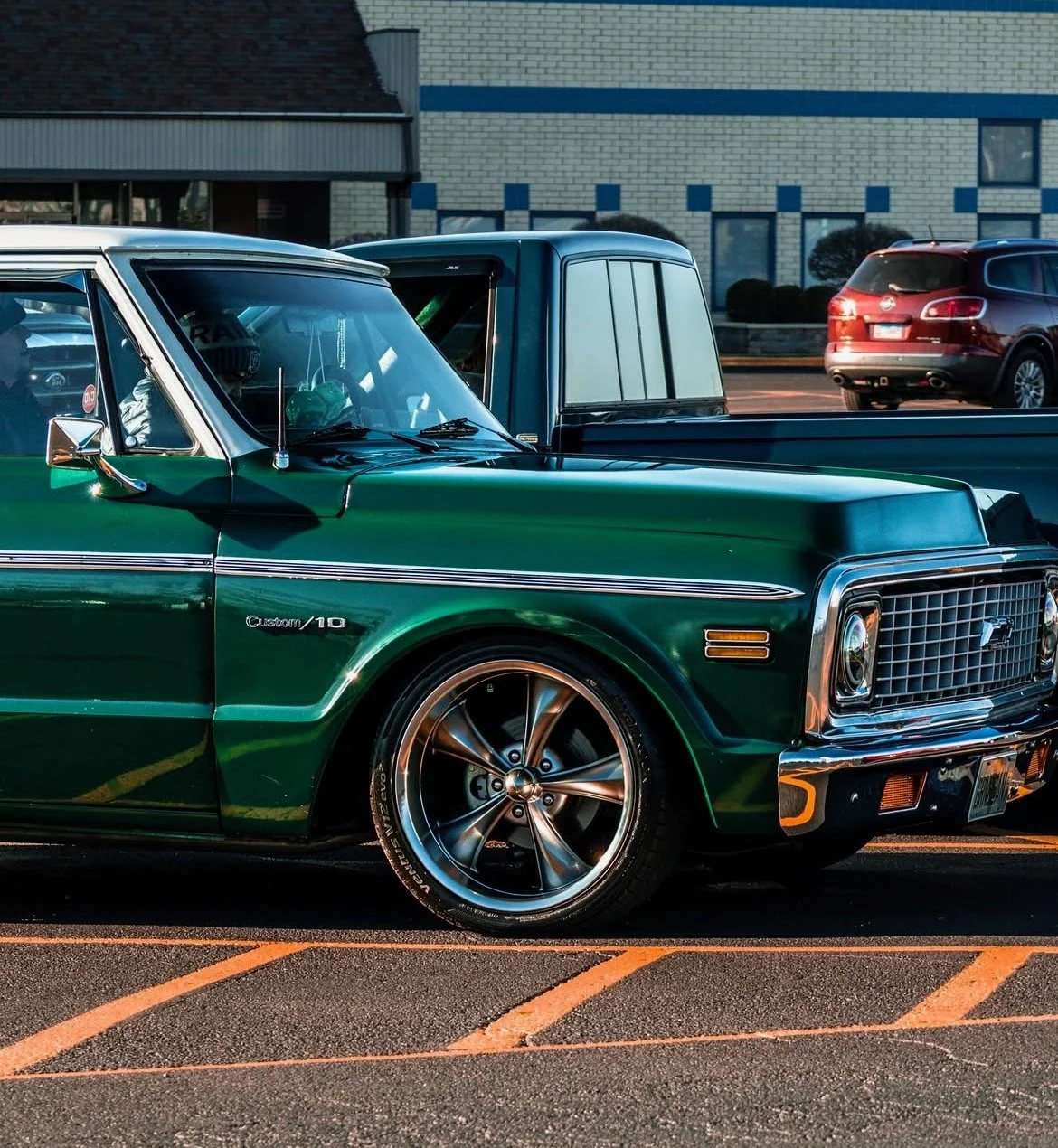 A close-up of a vintage green Chevrolet pickup truck parked in a lot, with modern vehicles in the background.