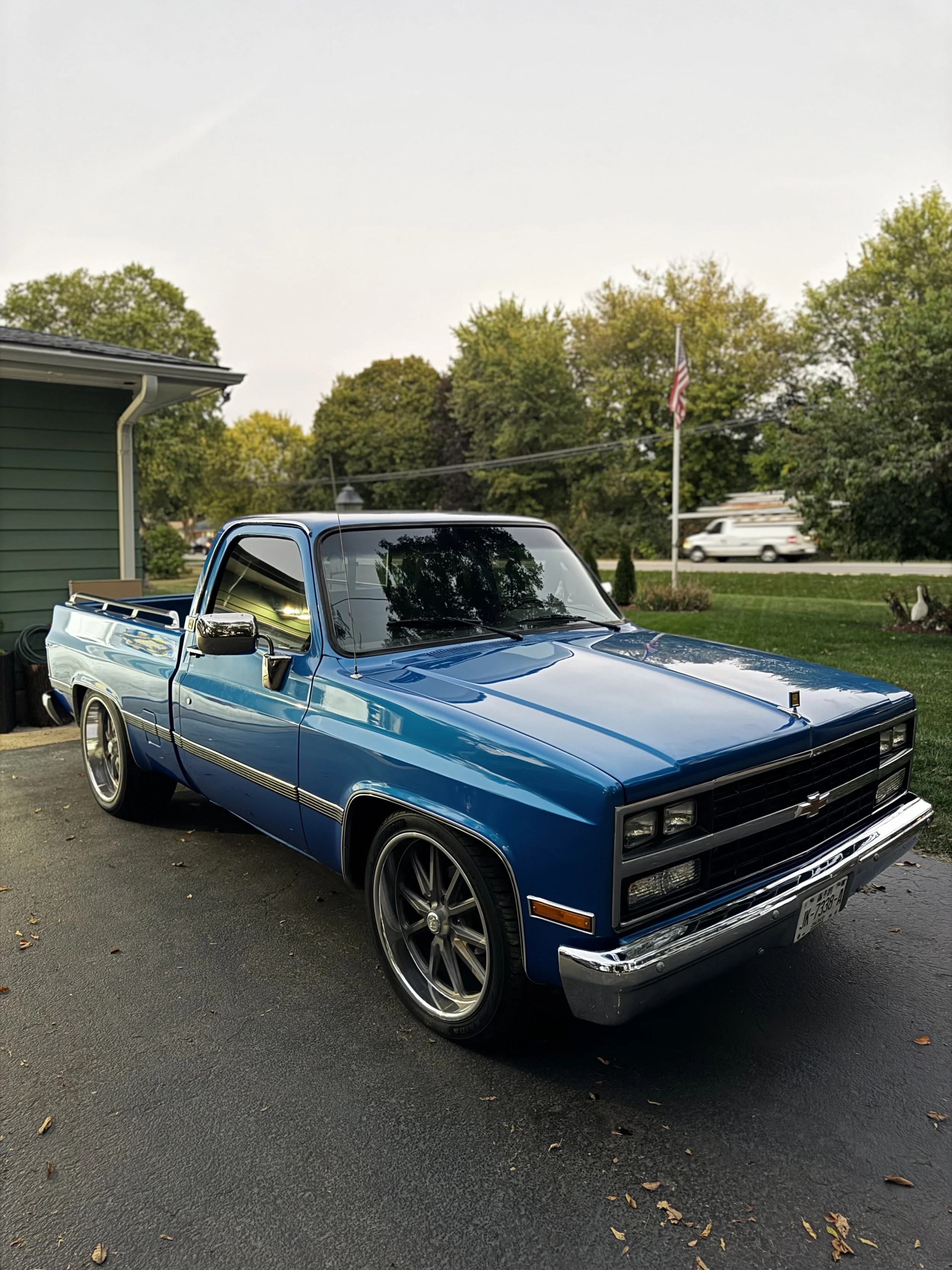 A blue vintage Chevrolet pickup truck parked on asphalt driveway near a green house with trees in the background.