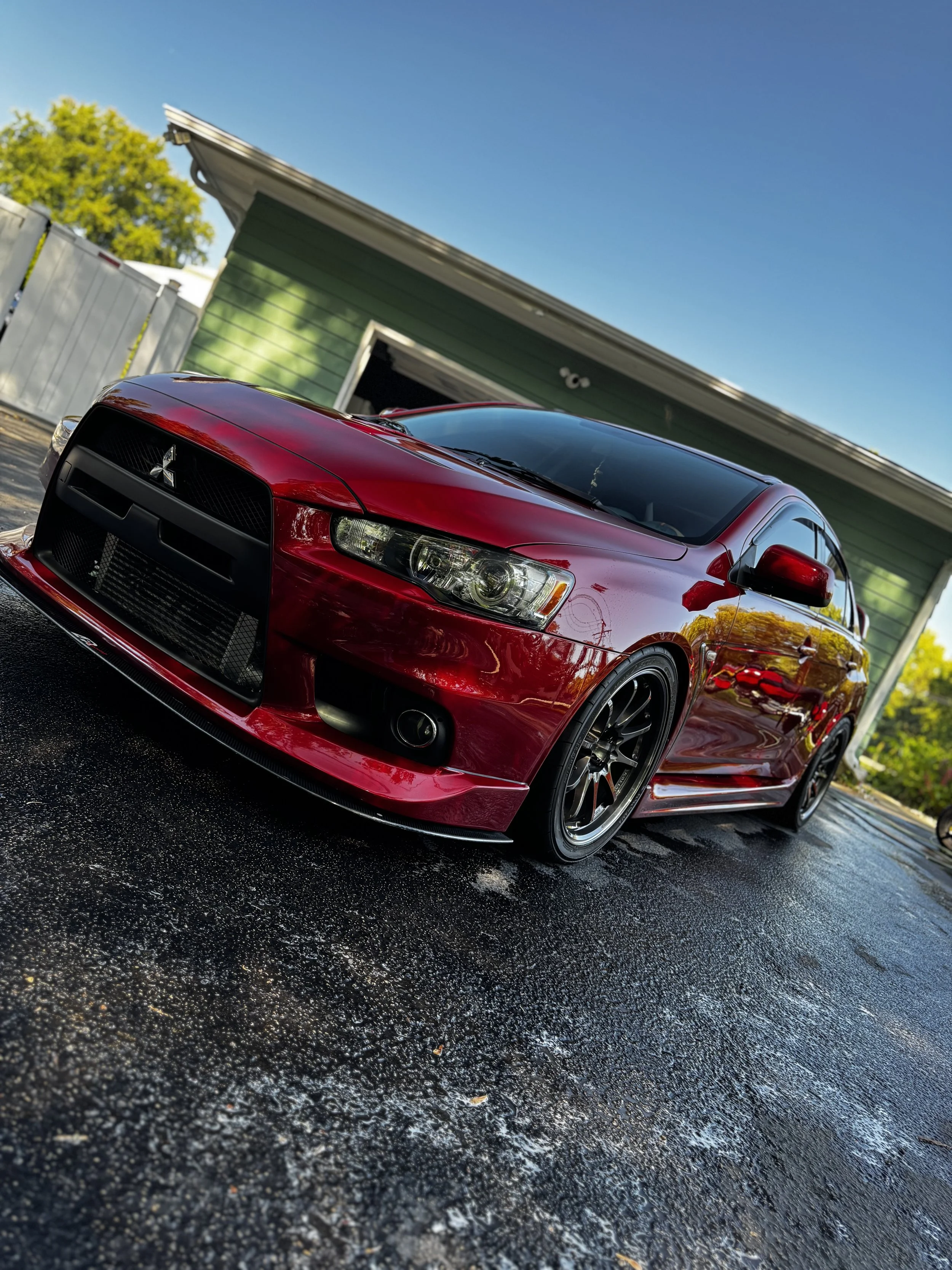 A red sports car parked on a wet driveway in front of a green house under a clear blue sky.
