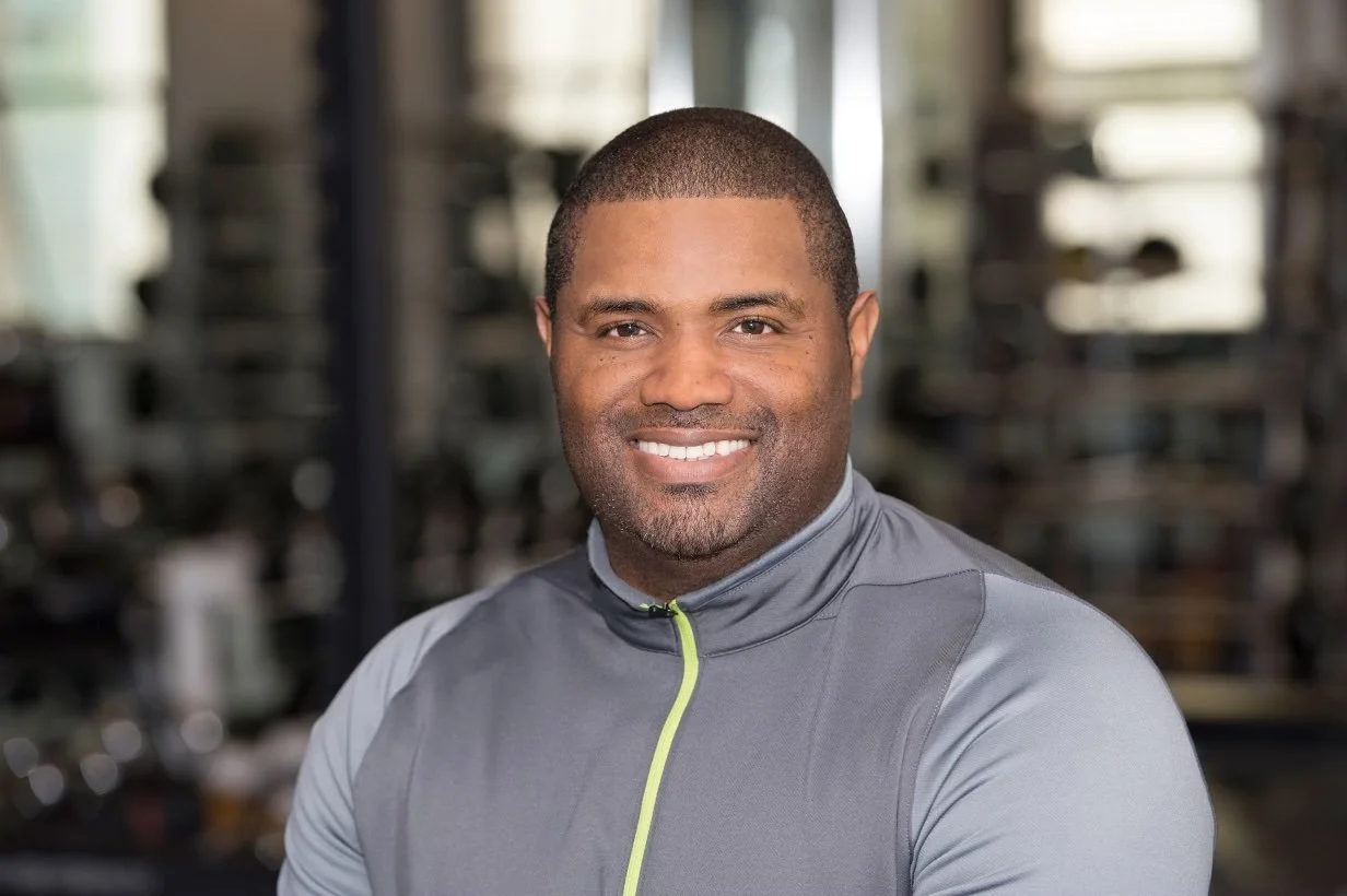 Smiling man in a grey zip-up jacket in a gym setting.