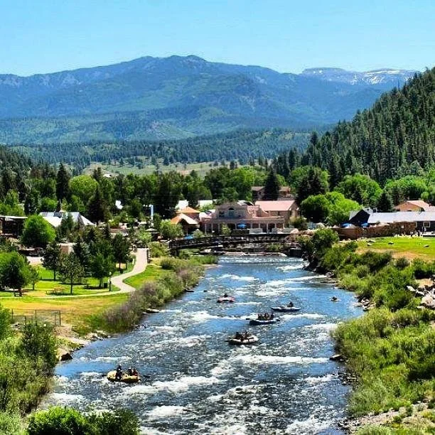 Scenic view of a river with boats, surrounded by green trees and a small town, with mountains in the background on a clear day.