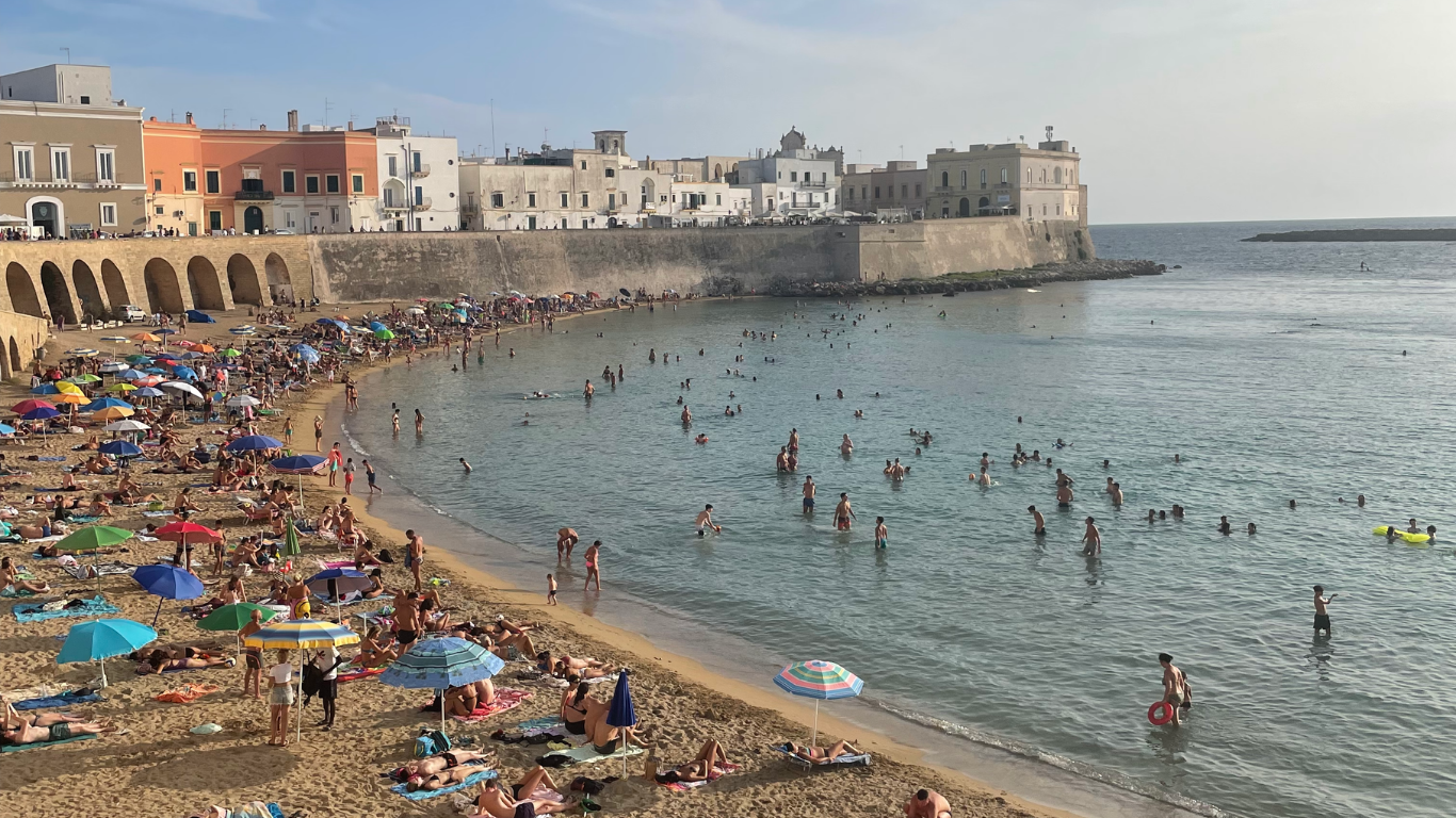 Crowded beach with colorful umbrellas, people sunbathing and swimming, historic buildings along the coast, calm sea under a partly cloudy sky.