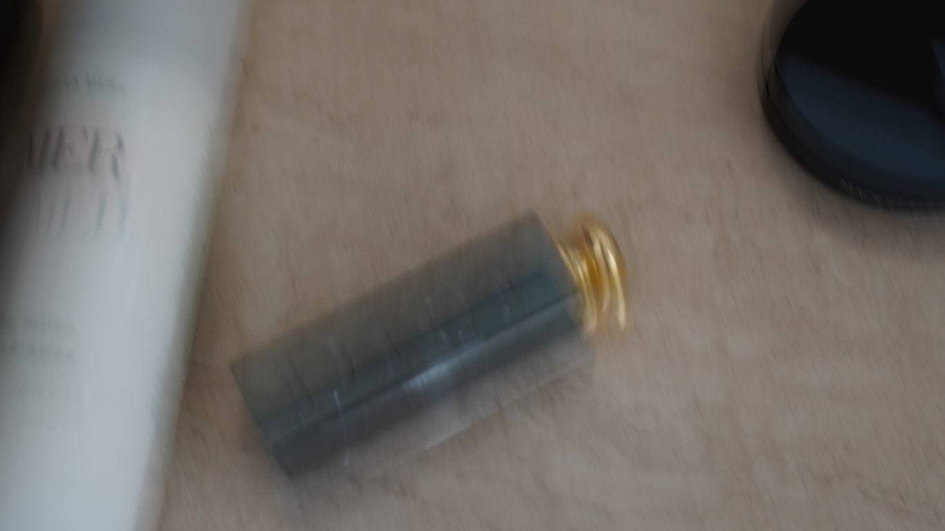 The image is blurry but it appears to contain a tube of some substance, a small object with a golden end, and part of a black and blue item on a light-colored surface.