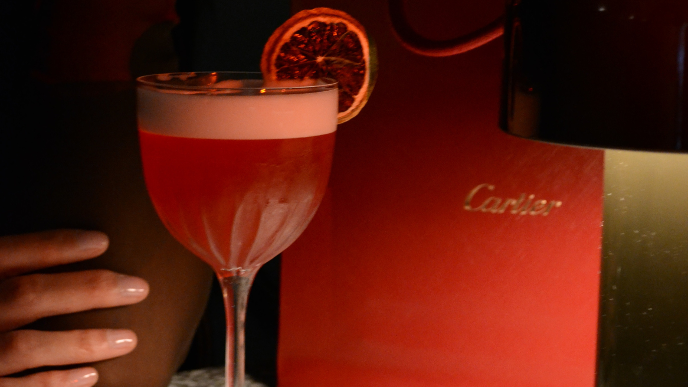 A hand holding a glass of pink cocktail with a dried citrus slice garnish, next to a red Cartier shopping bag.