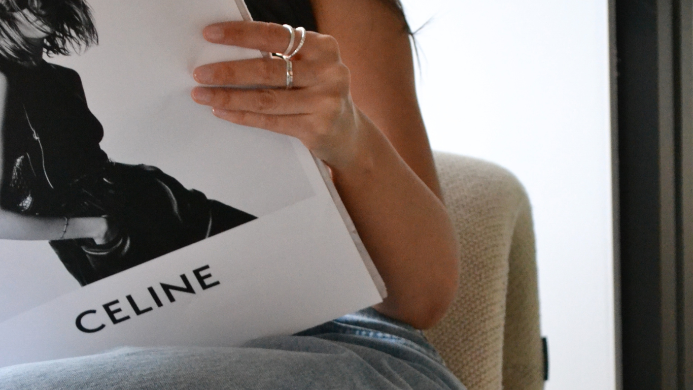 Person holding a Celine magazine, sitting on a beige armchair, with their arm and hand with rings visible, wearing a dark sleeveless top and denim shorts.