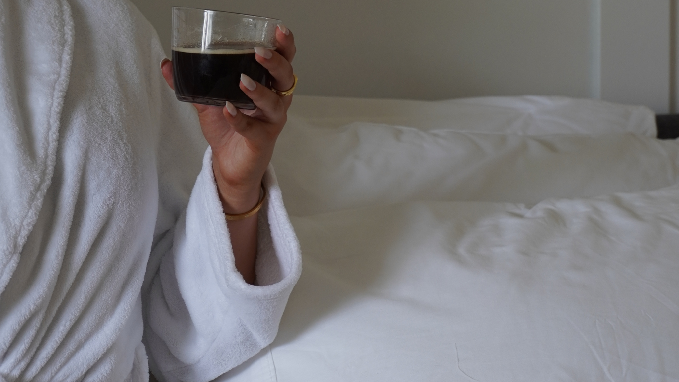 Person in a white bathrobe holding a glass of dark beverage in bed with white bedding.
