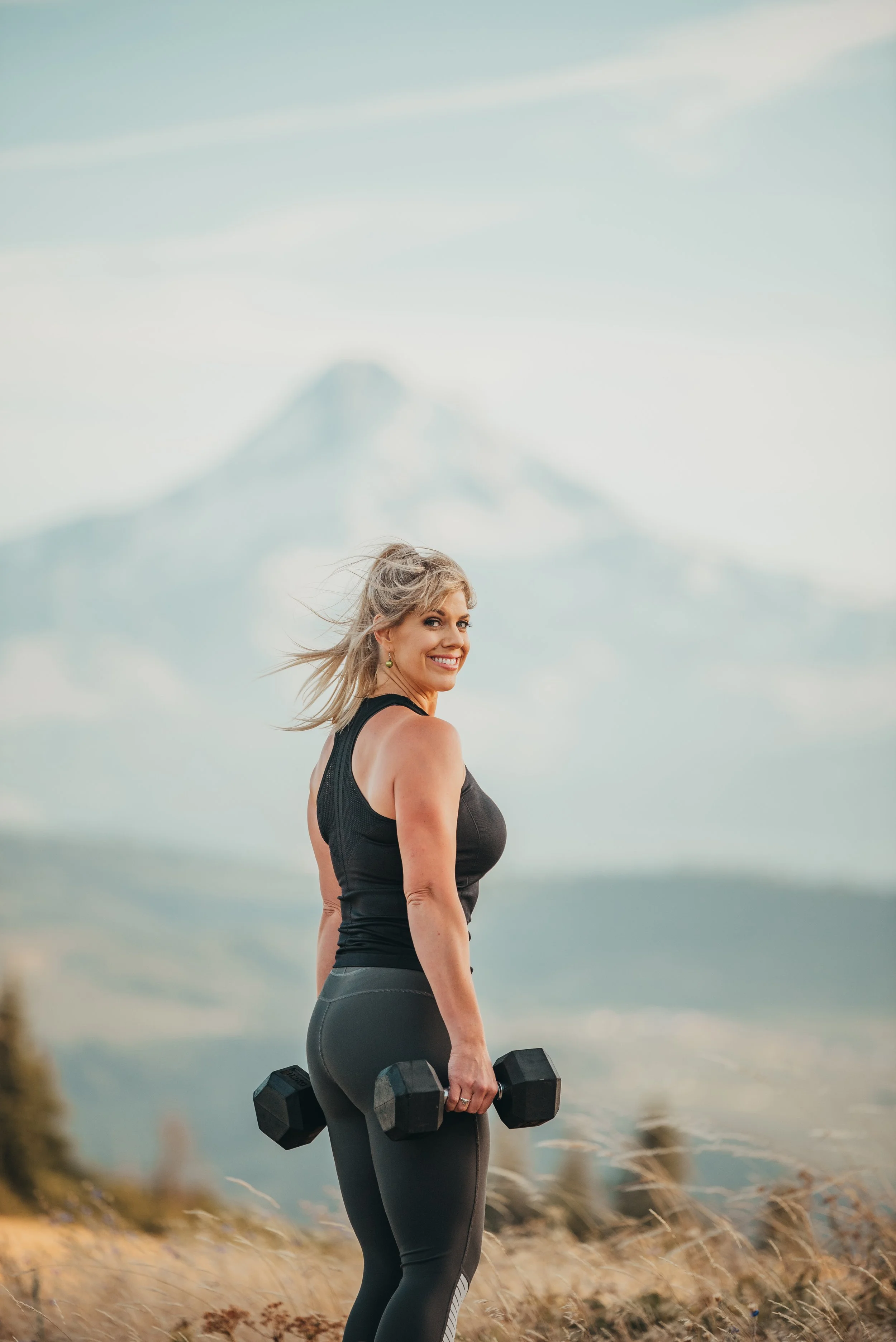 Coach Laura smiles looking back at the camera, with weights in hand and Mt. Hood in the background