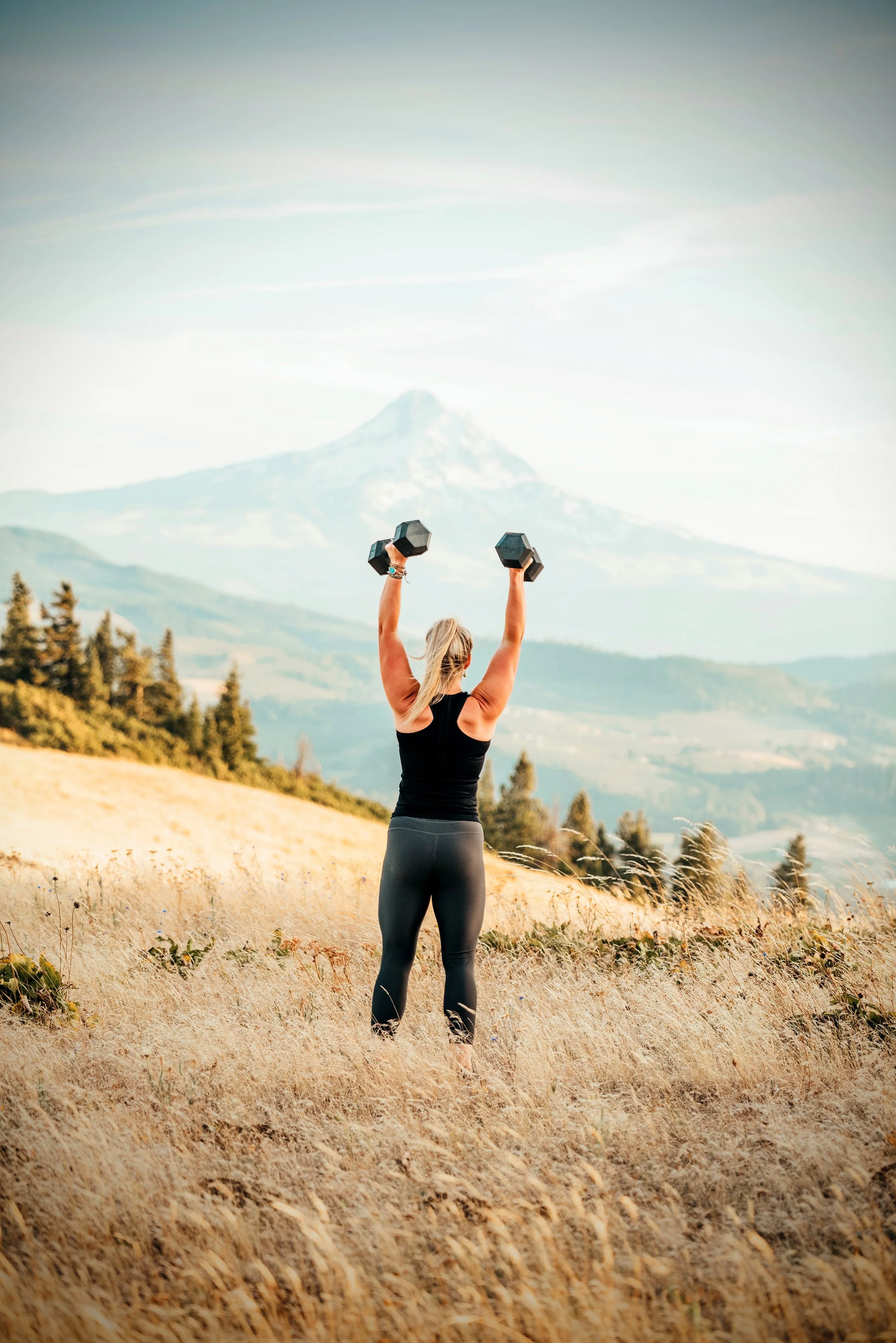 Coach Laura is holding hand weights above her head while facing Mt. Hood