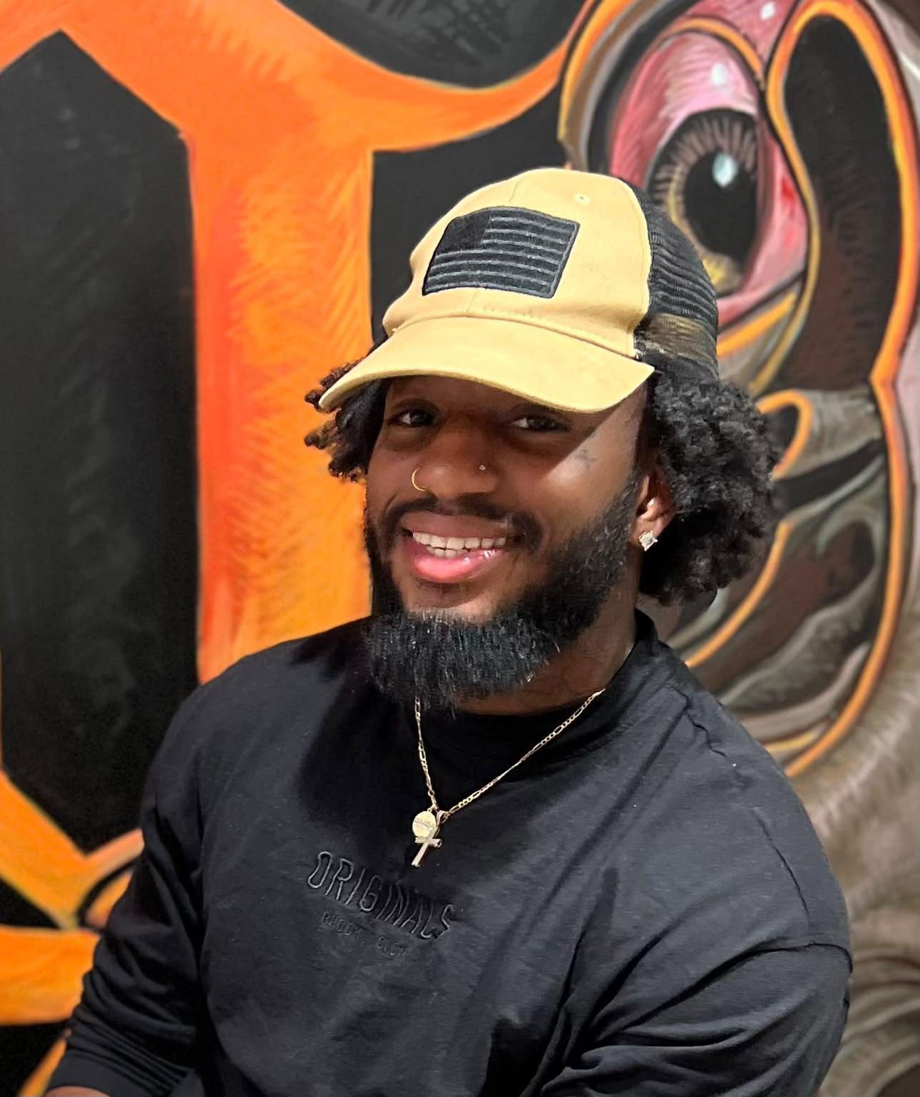 A smiling man with a beard and mustache wearing a beige cap with a black patch, a nose piercing, earrings, a chain necklace, and a black shirt, posing in front of colorful, abstract artwork.