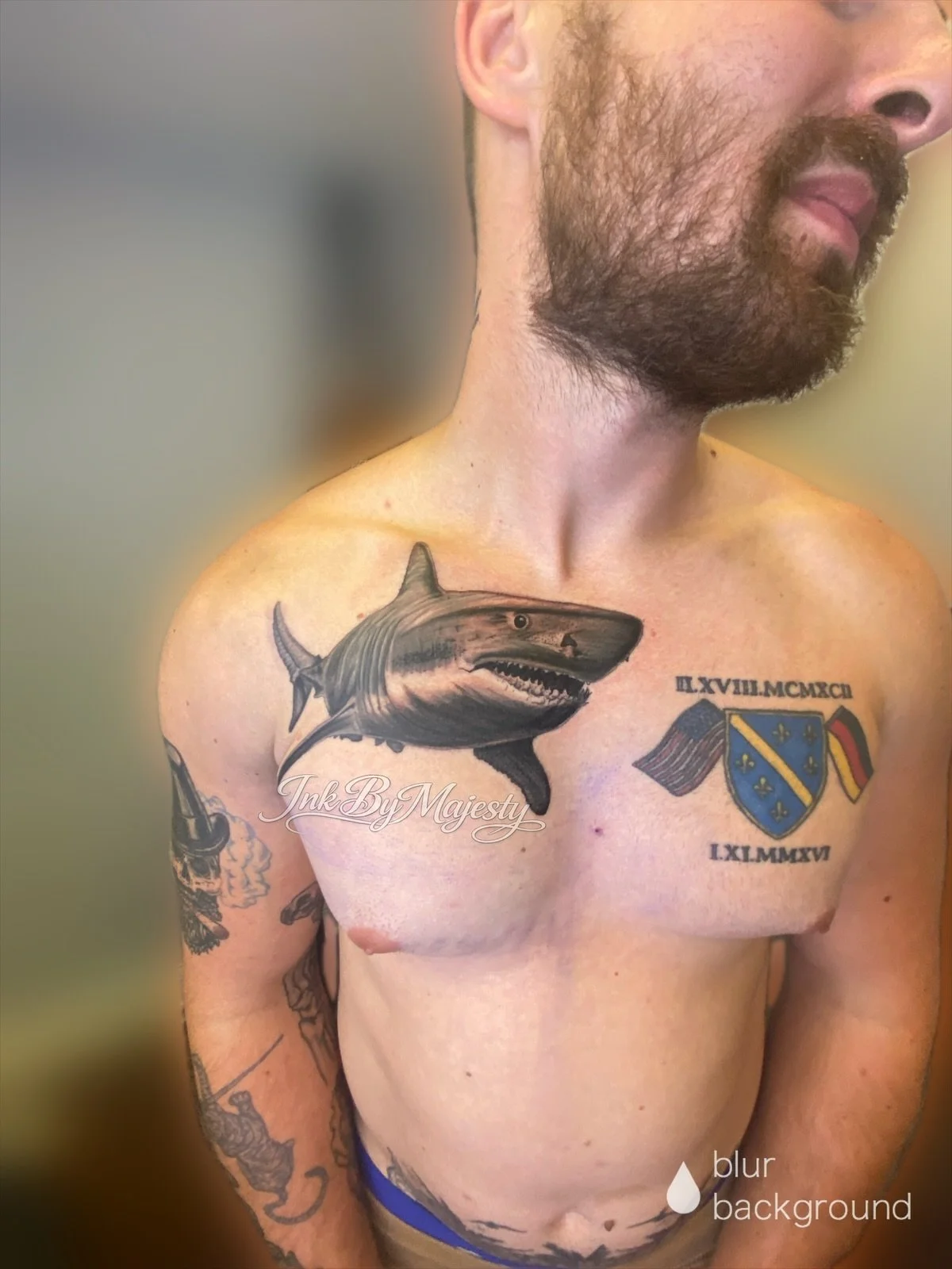 A shirtless man with tattoos on his chest and arms, including a shark tattoo on his left shoulder and a shield with flags on his right chest, standing against a blurred background.