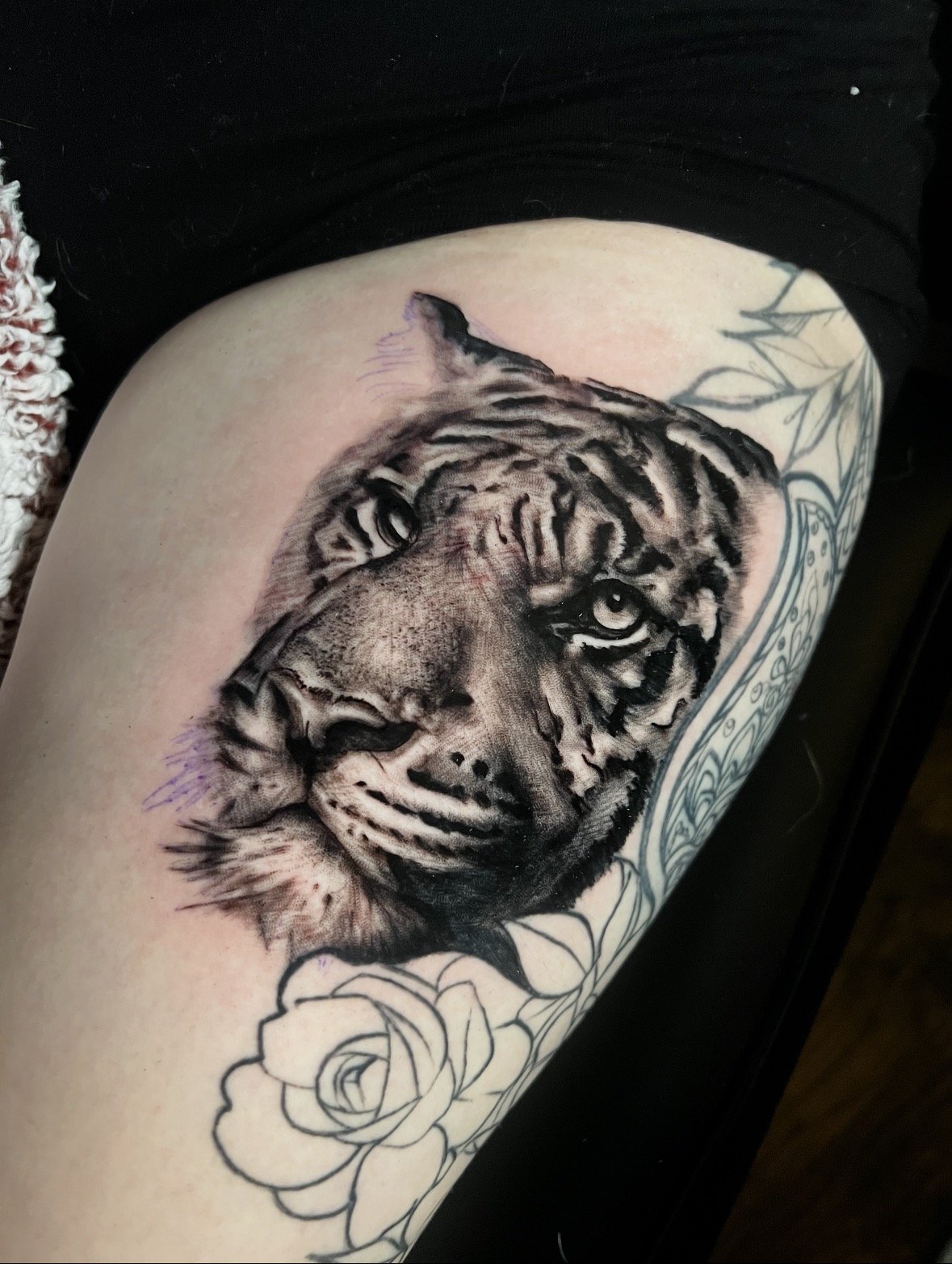 Tattoo of a detailed tiger's face with black and gray shading on someone's upper arm, with some floral line art around it.