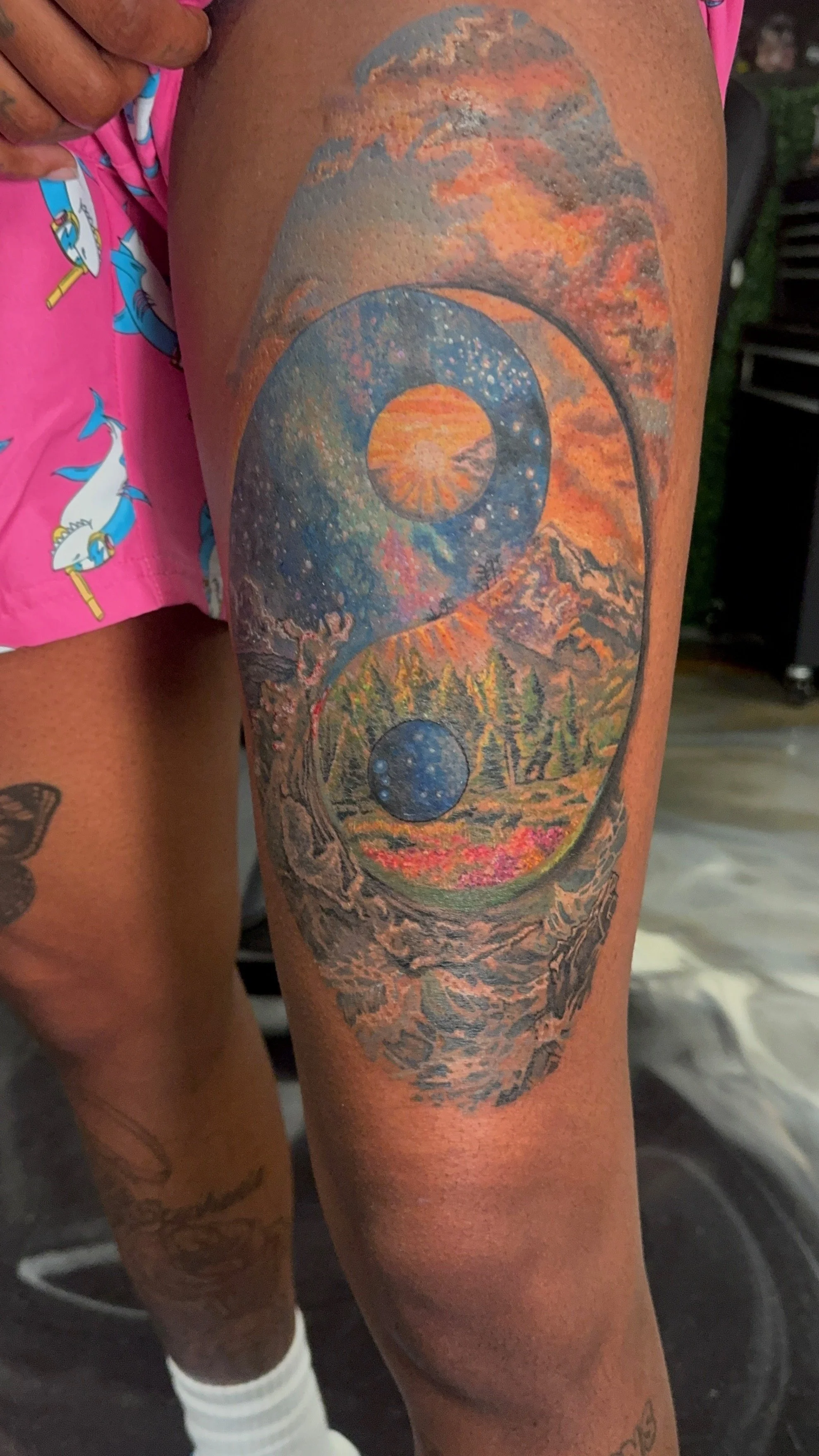 Tattoo of a cosmic landscape featuring moons, mountains, trees, and a sunset sky on a person's thigh.