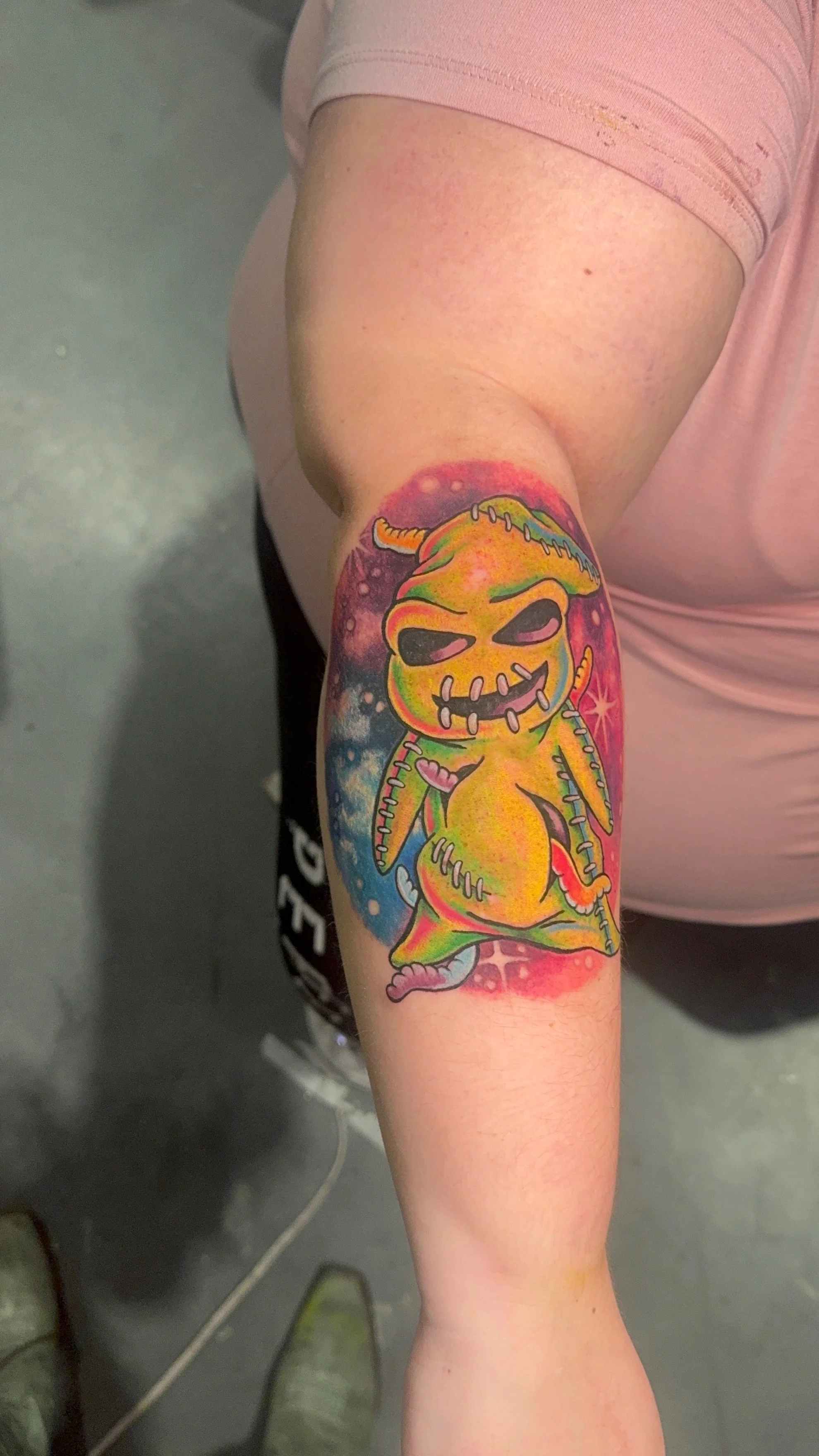 Colorful tattoo of a cartoonish creature with stitched features, dark eyes, and a mischievous smile on a person's arm.