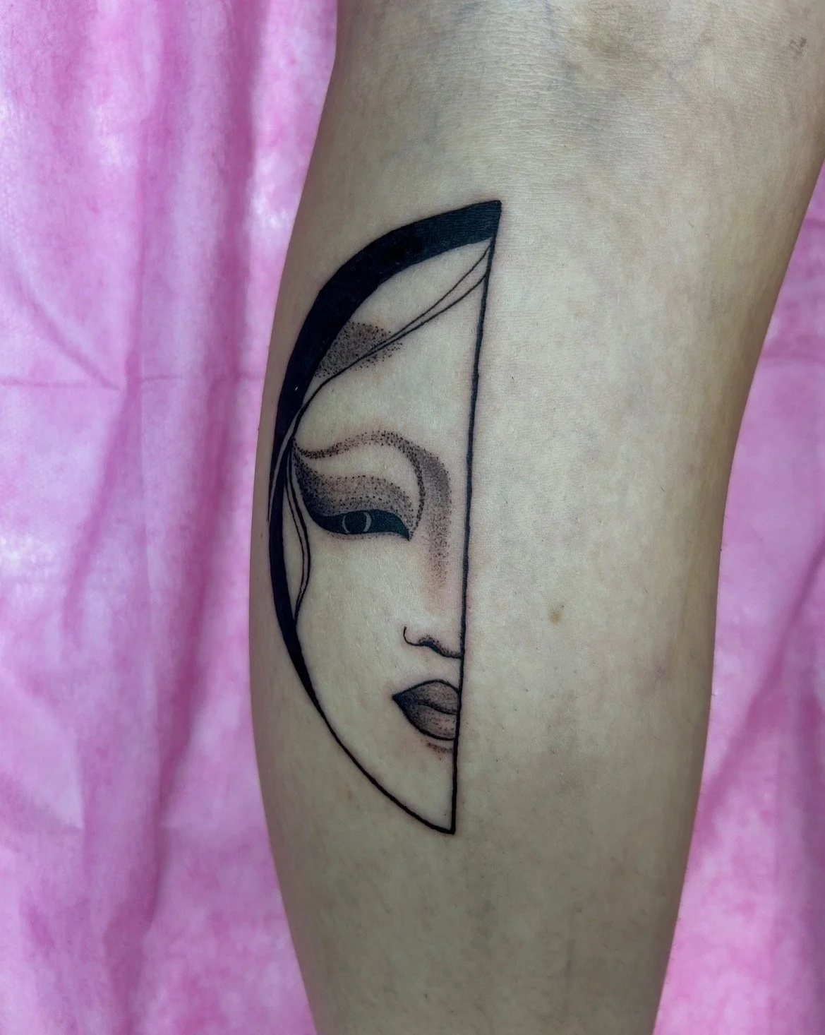 Tattoo of a stylized female face with half of it inside a geometric arc, on a person's arm, with a pink fabric background.